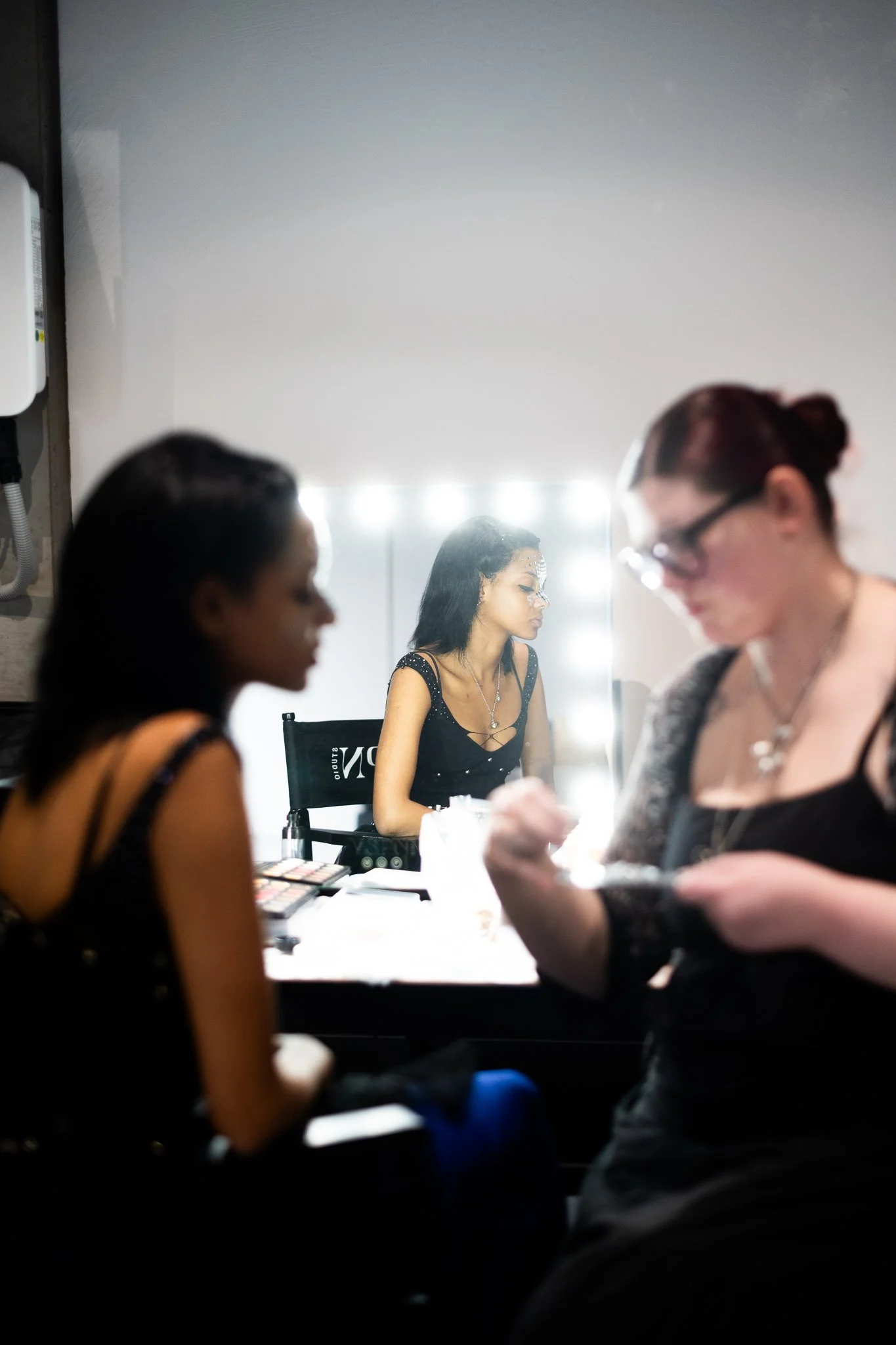 Three women are in a room, with one woman sitting at a table applying makeup, while the other two women are standing nearby. The scene is illuminated by bright lights around a mirror in the background.