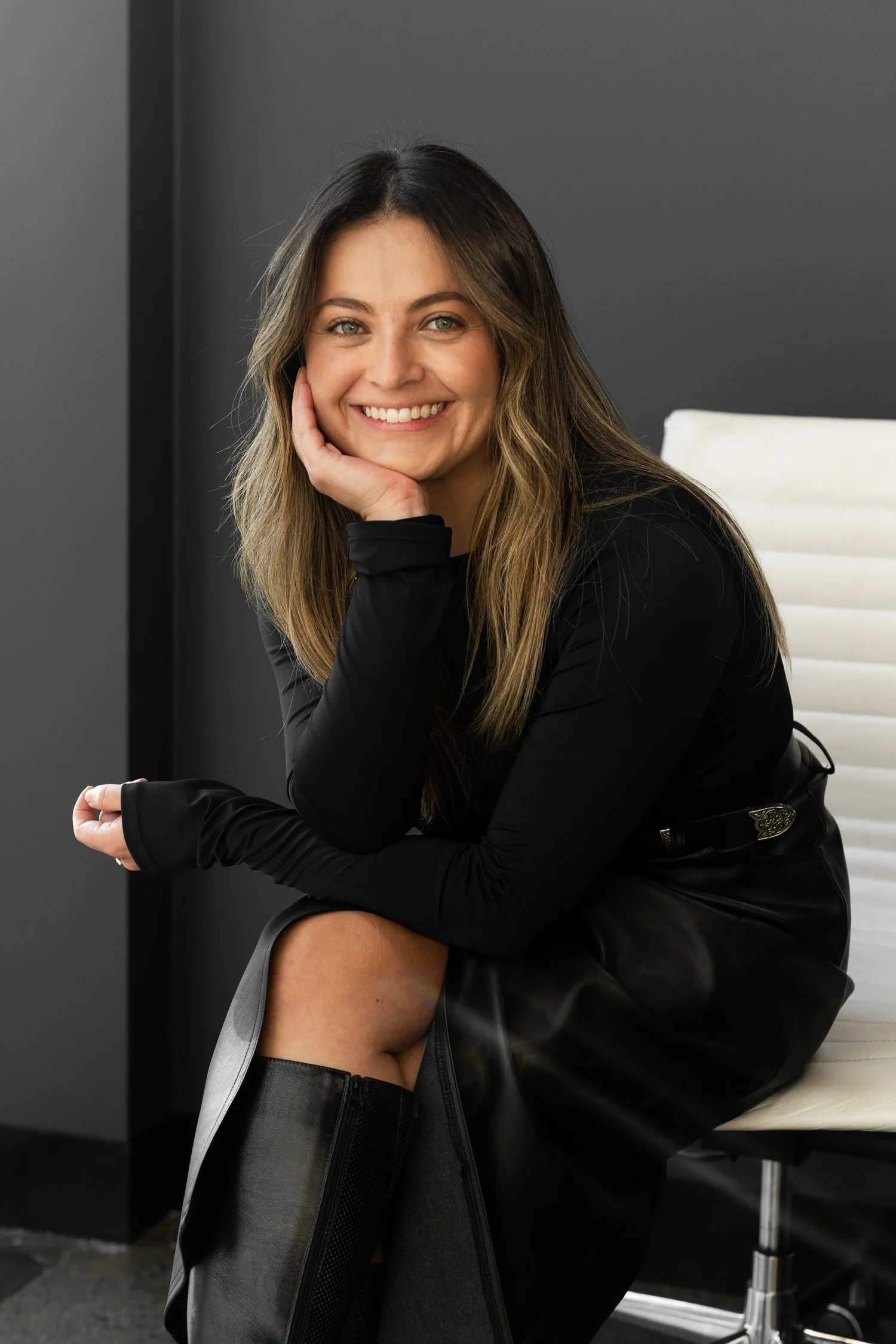 A woman with long light brown hair and green eyes, smiling and resting her chin on her hand, seated on a white chair in a modern office with dark gray walls.