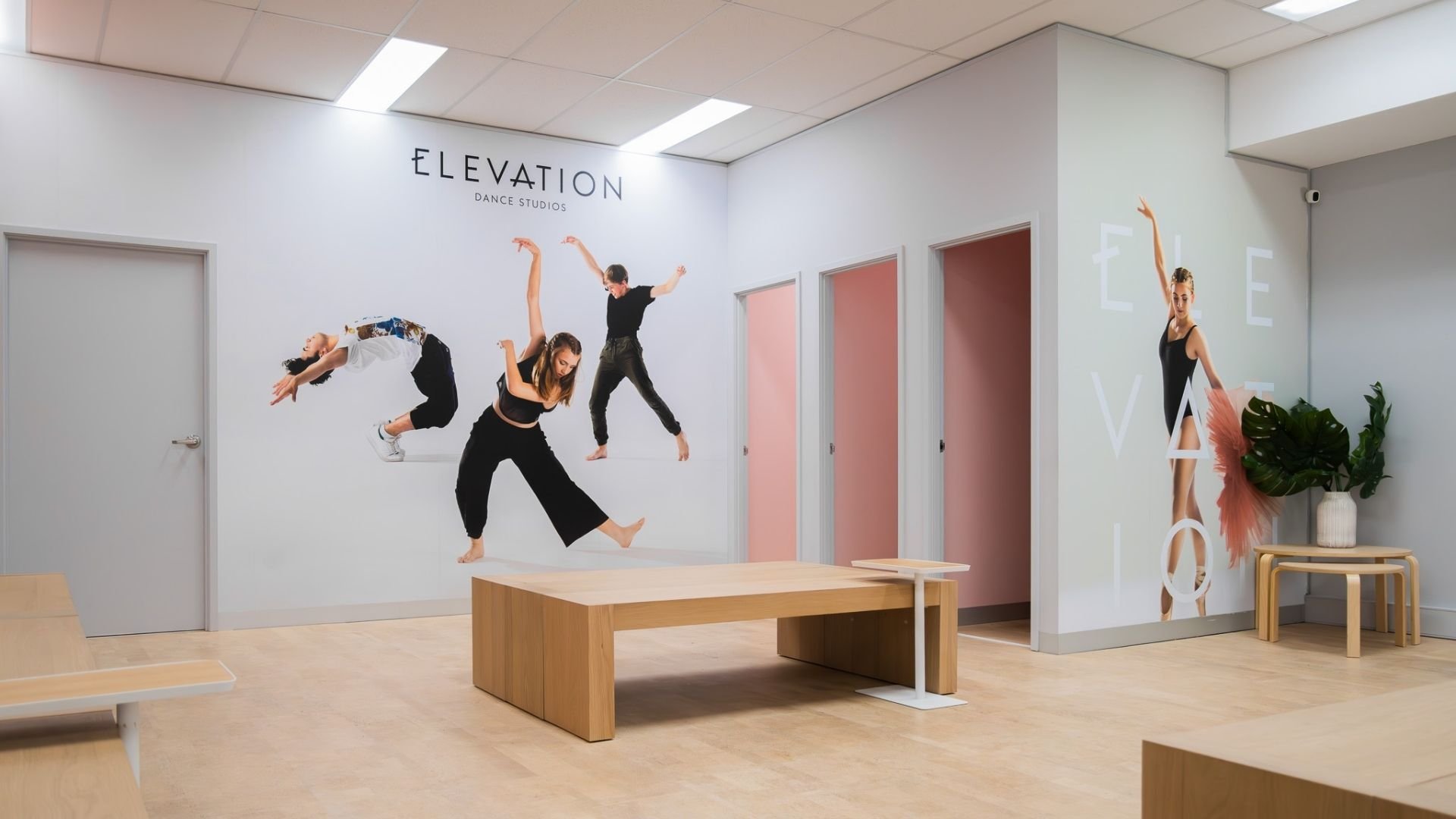 Dance studio interior with a mural of four dancers in various dynamic poses on the white wall behind, with the words 'ELEVATION DANCE STUDIOS' at the top, and a life-sized poster of a ballerina on the adjacent wall, decorated with green plants and mo