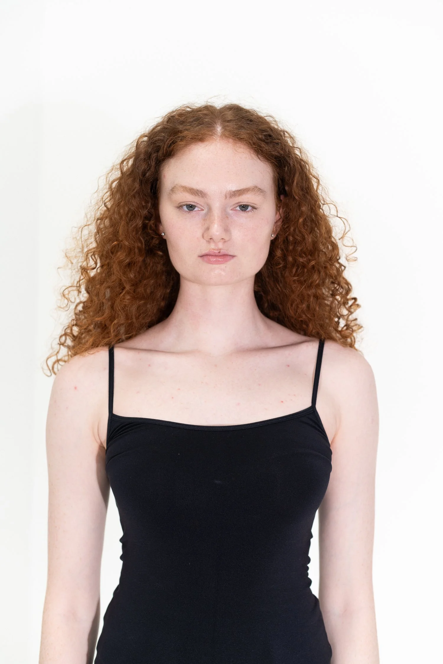 Portrait of a young woman with curly red hair, wearing a black spaghetti strap top, standing against a plain off-white background.