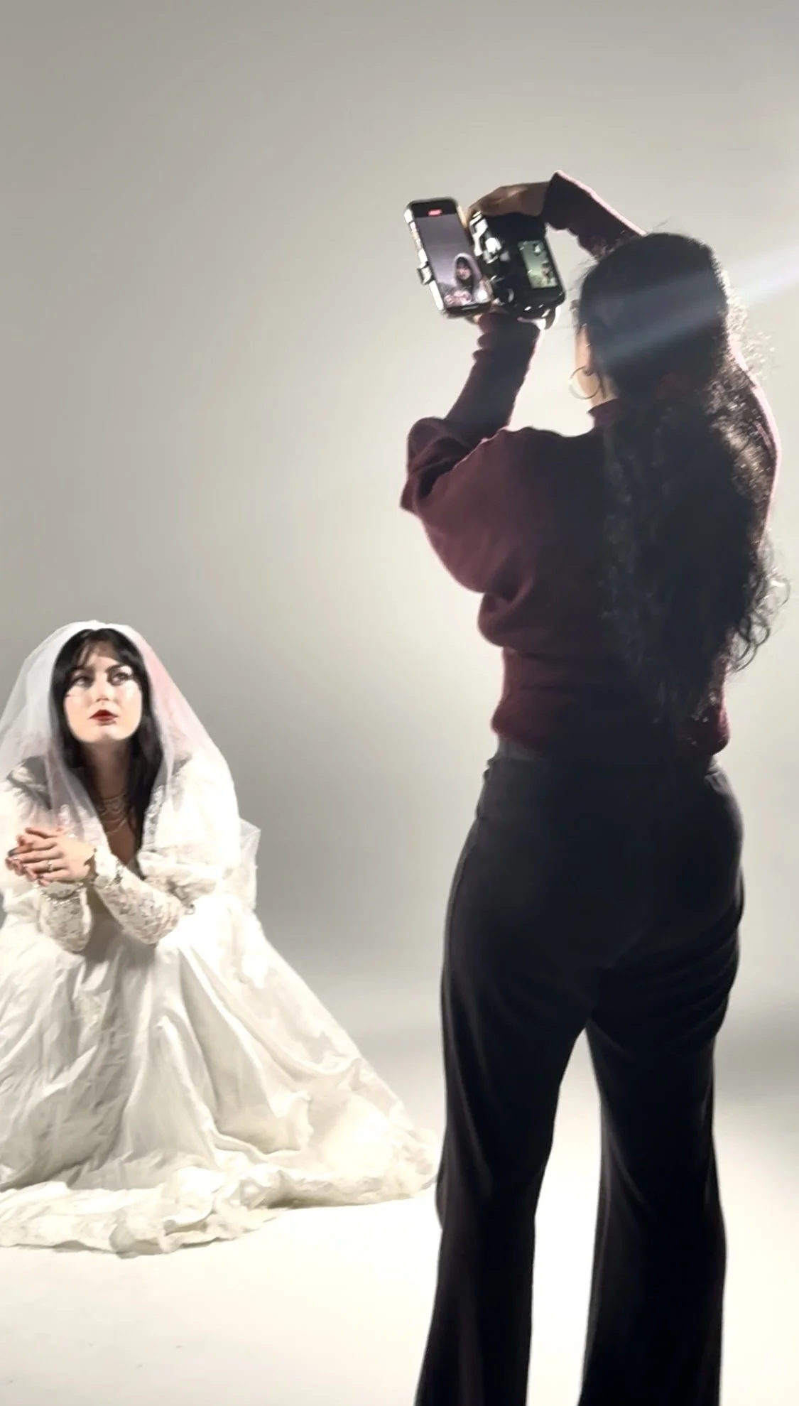 A woman dressed in white with a veil, sitting on the floor, posing for a photo in front of a plain gray background while a photographer takes her picture. Photography session with models. Talent sourcing