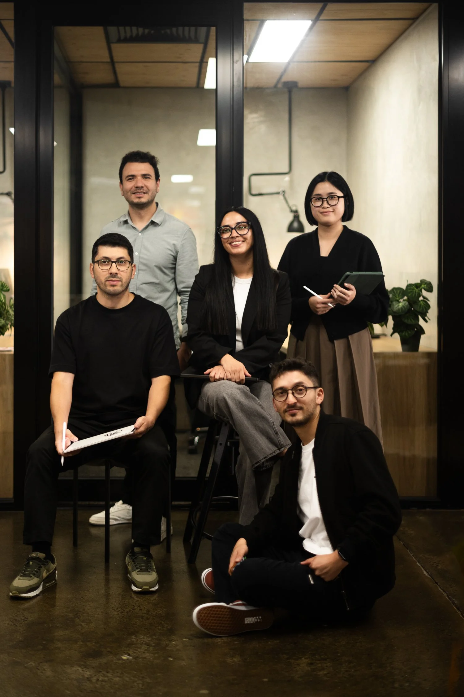 A group of five diverse young adults posing inside a modern office space with large glass windows and plants, some sitting and others standing, smiling at the camera.
