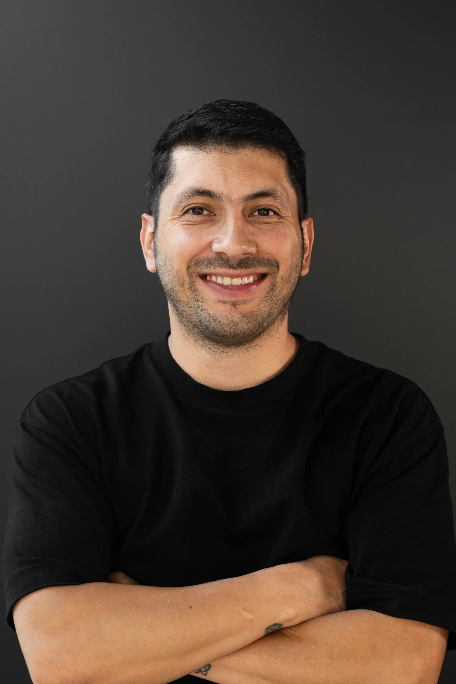 A smiling man with dark hair and a beard, dressed in a black shirt, with arms crossed, against a dark background.