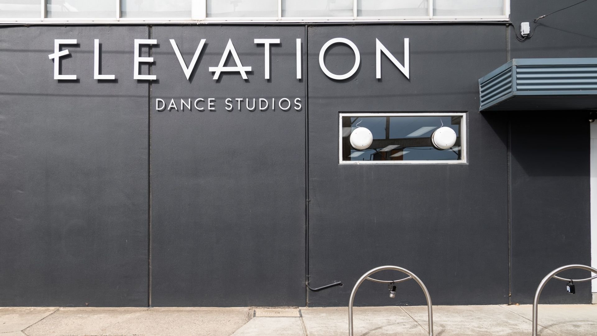 Exterior view of a dark gray building with a sign that reads 'ELEVATION DANCE STUDIOS' in large white letters. There is a small rectangular window with two circular white lights inside and a metallic awning to the right.