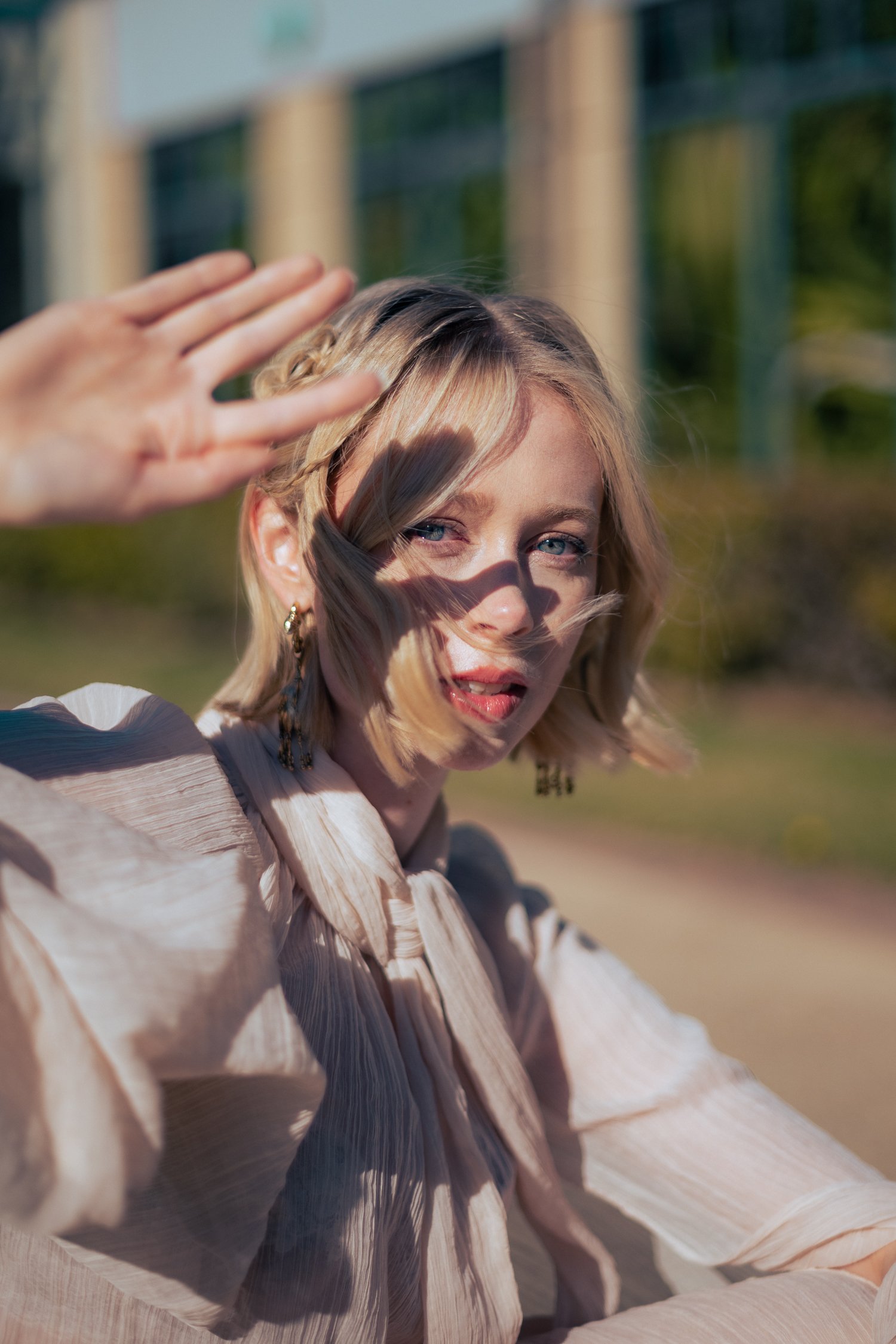 A woman with blonde hair and blue eyes outdoors, shading her face with her hand, wearing a beige striped blouse.