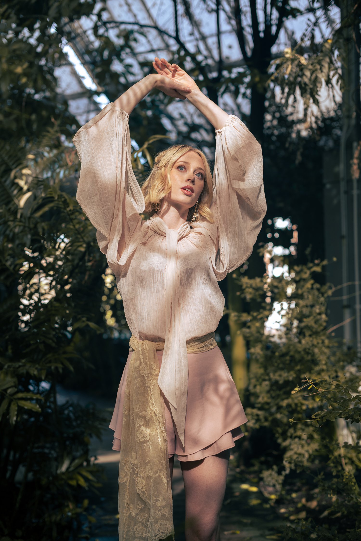 A woman with blonde hair wearing a light pink dress with lace and tulle details, standing among green foliage, with her arms raised above her head in a dance pose.