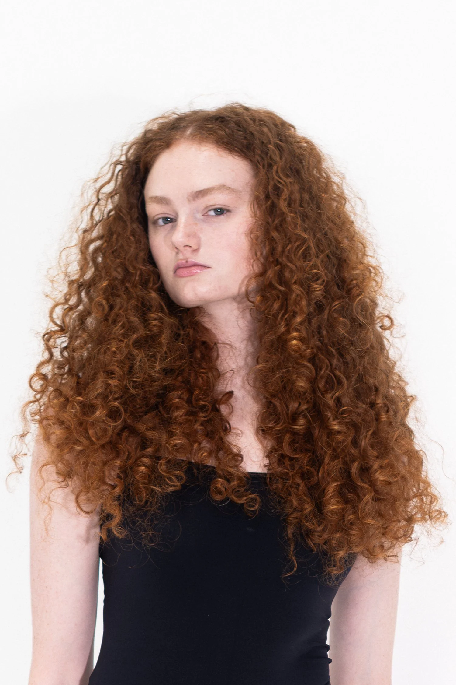 Close-up of a woman with long, curly red hair, light skin, and blue eyes wearing a black sleeveless top against a plain white background.