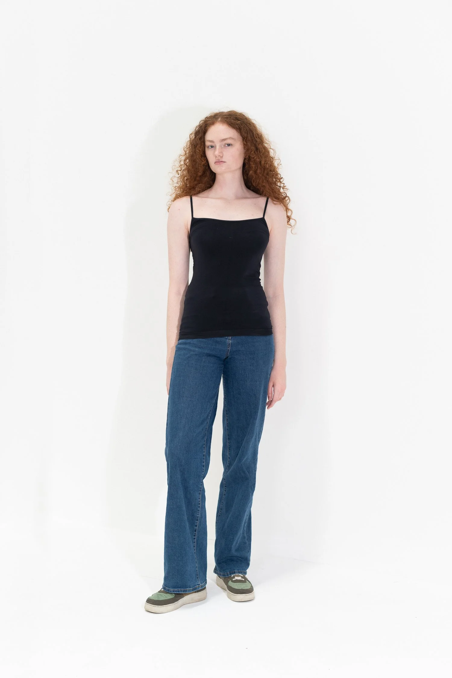 A young woman with long, curly red hair and fair skin standing against a white background. She is wearing a black spaghetti strap top, blue jeans, and sneakers.