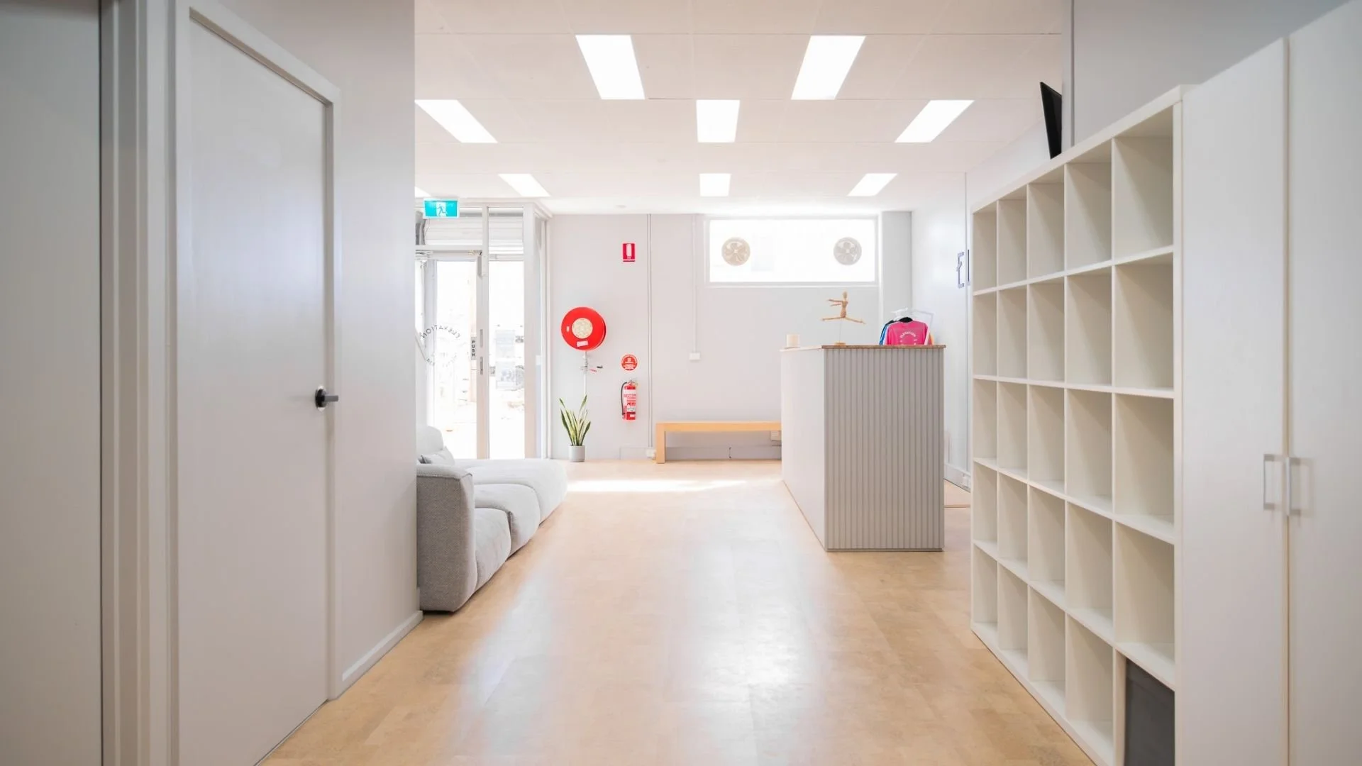 Bright, minimalistic reception area with a white couch, potted plant, and a large white shelving unit on the right. The space has white walls, a light wooden floor, and ceiling lights. There are fire safety signs and a door with glass panels at the b