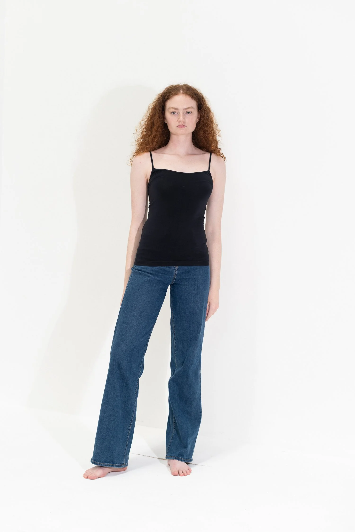 Young woman with curly red hair standing barefoot against a white background, wearing a black spaghetti strap top and blue jeans.