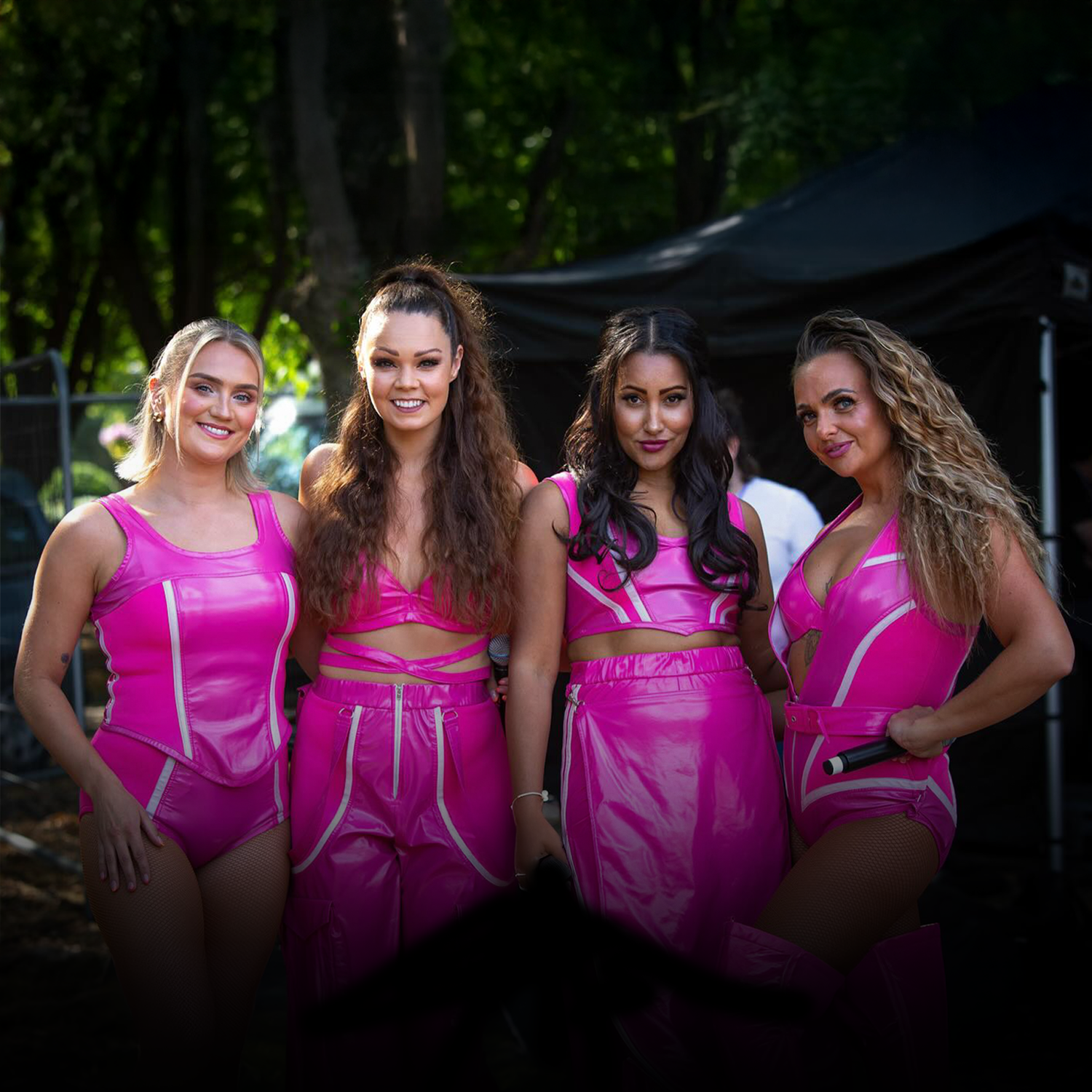 Four women in pink costumes standing outdoors on a sunny day, smiling at the camera.
