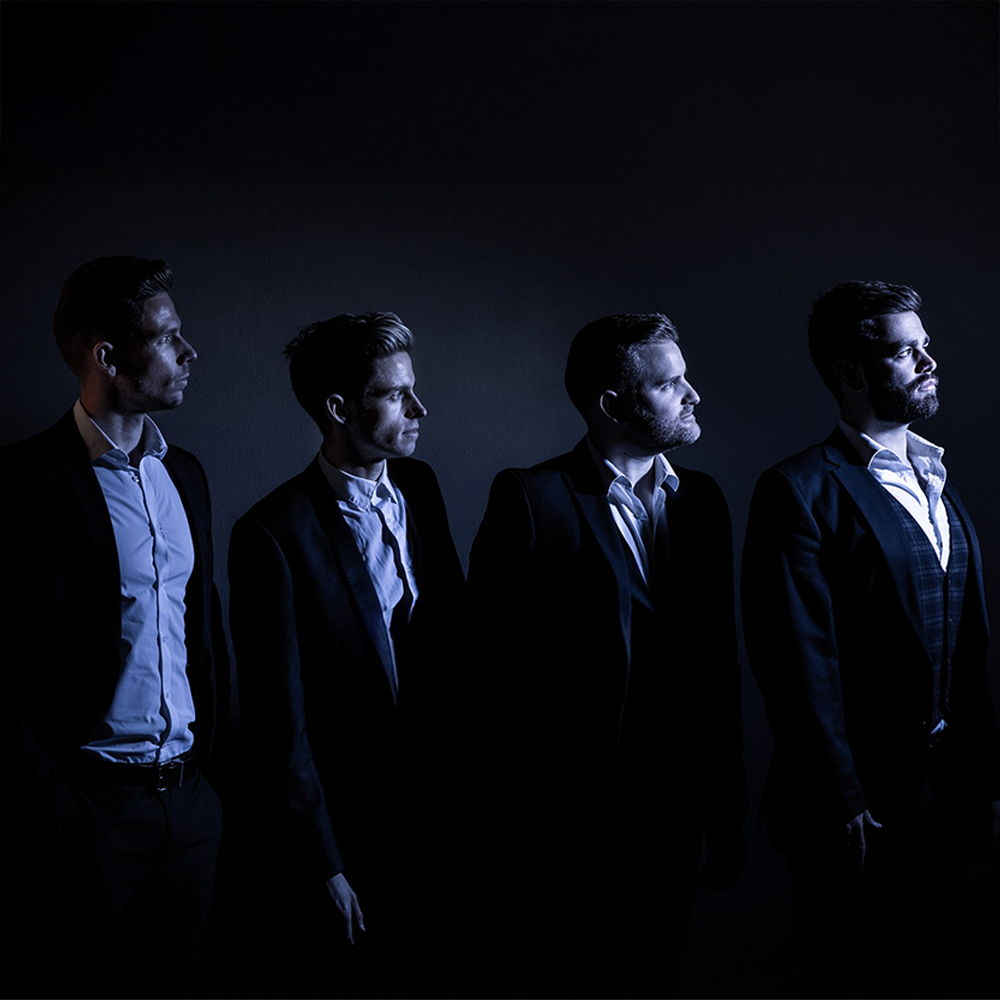 Four men in suits and white shirts, standing in profile against a dark background, with side lighting highlighting their faces and neutral expressions.