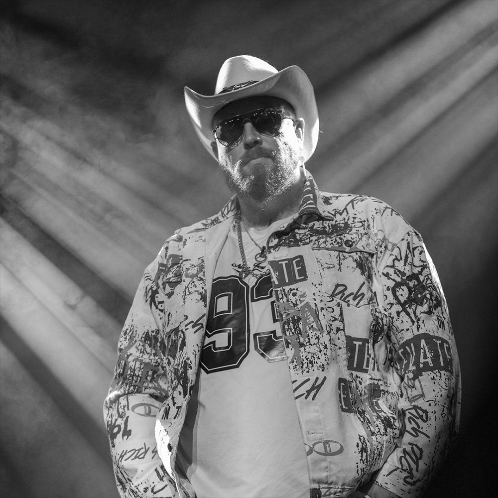 A man wearing a cowboy hat, sunglasses, a patterned jacket, and a T-shirt with the number 94, standing in a spotlight with beams of light radiating behind him in a black-and-white photograph.