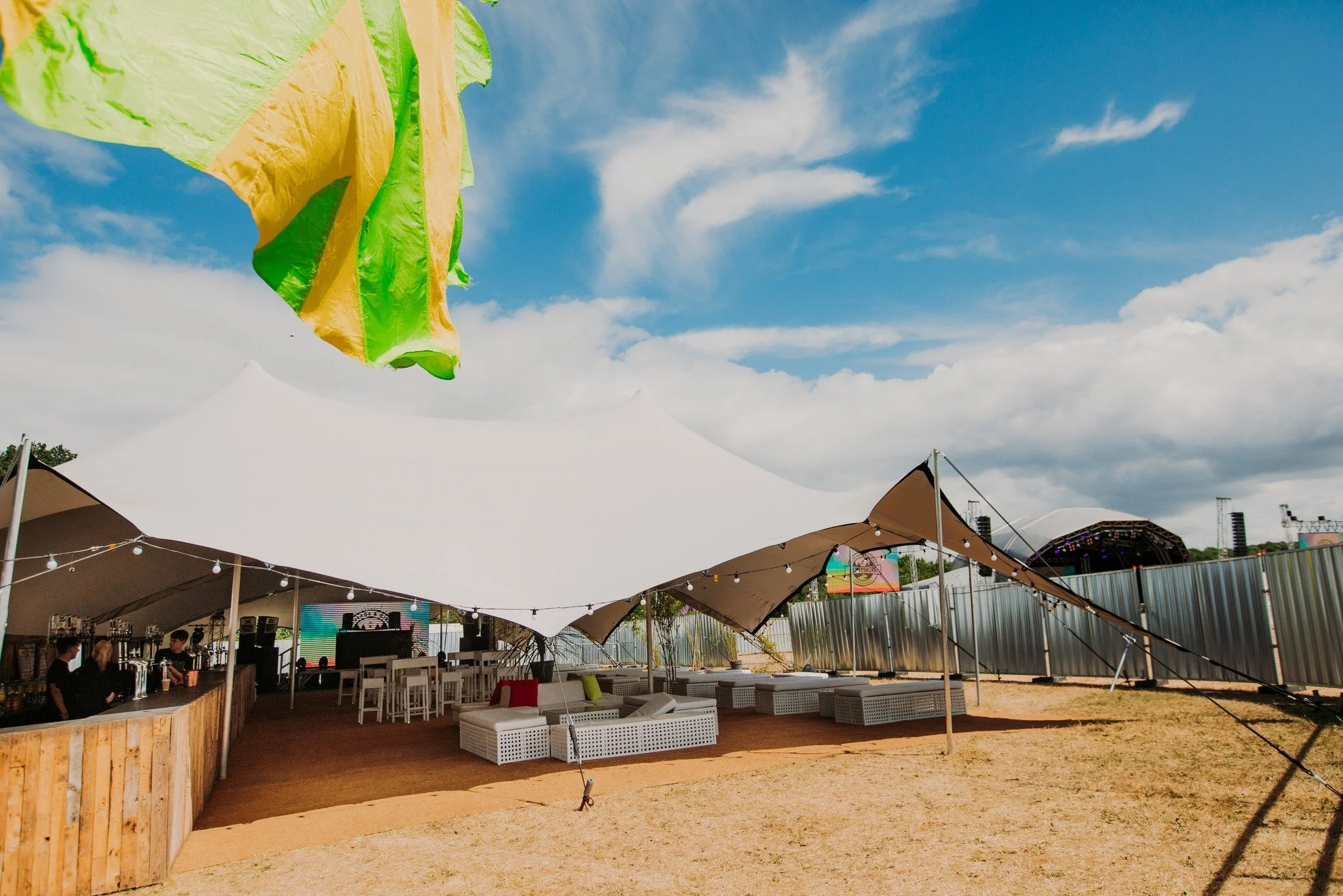 An outdoor event setup under a large white tent with string lights, beige wicker seating, and a bar area, with a stage in the background. A colorful flag or banner is in the upper left corner, and the sky is partly cloudy with blue sky visible.