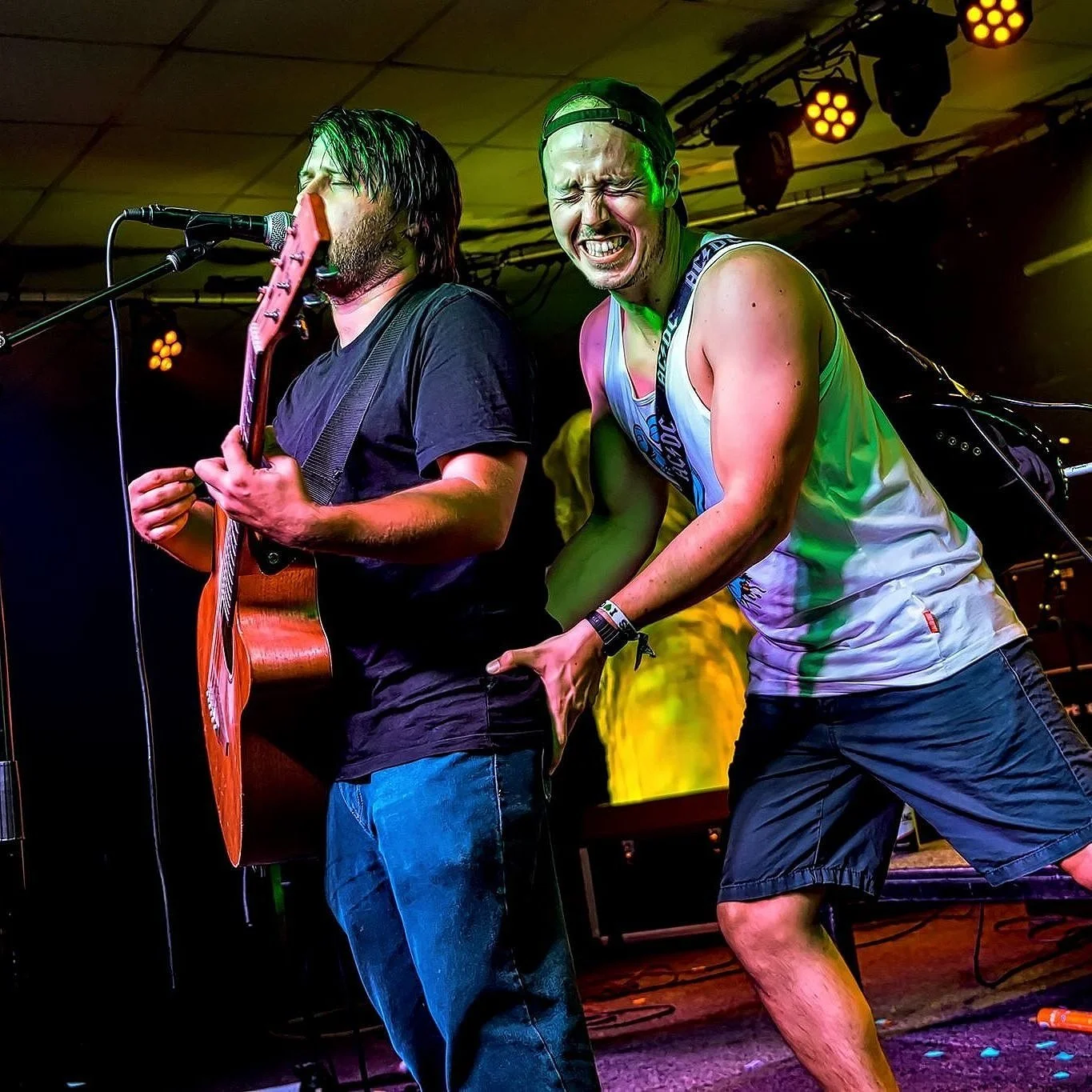 Two men performing live on stage, one playing guitar and singing into a microphone, the other dancing and showing an energetic expression, with vibrant stage lighting and equipment in the background.