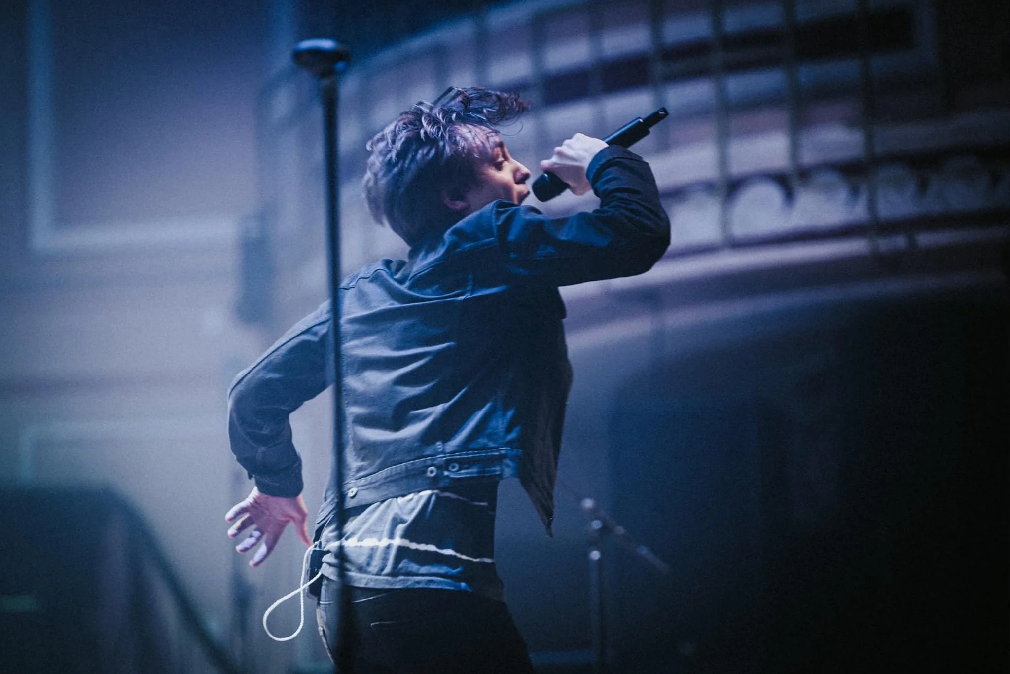 Performer singing passionately into a microphone on stage, wearing a black jacket and jeans, with a dark background and stage lighting.