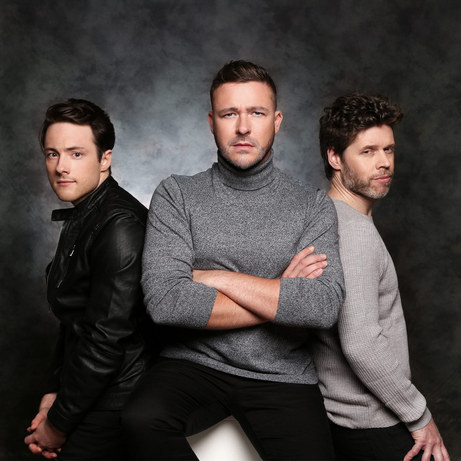 Three men posing against a dark, textured background. The man in the center is wearing a gray turtleneck sweater with arms crossed. The man on the left is in a black leather jacket, and the man on the right is in a light gray sweater. They are looking toward the camera.