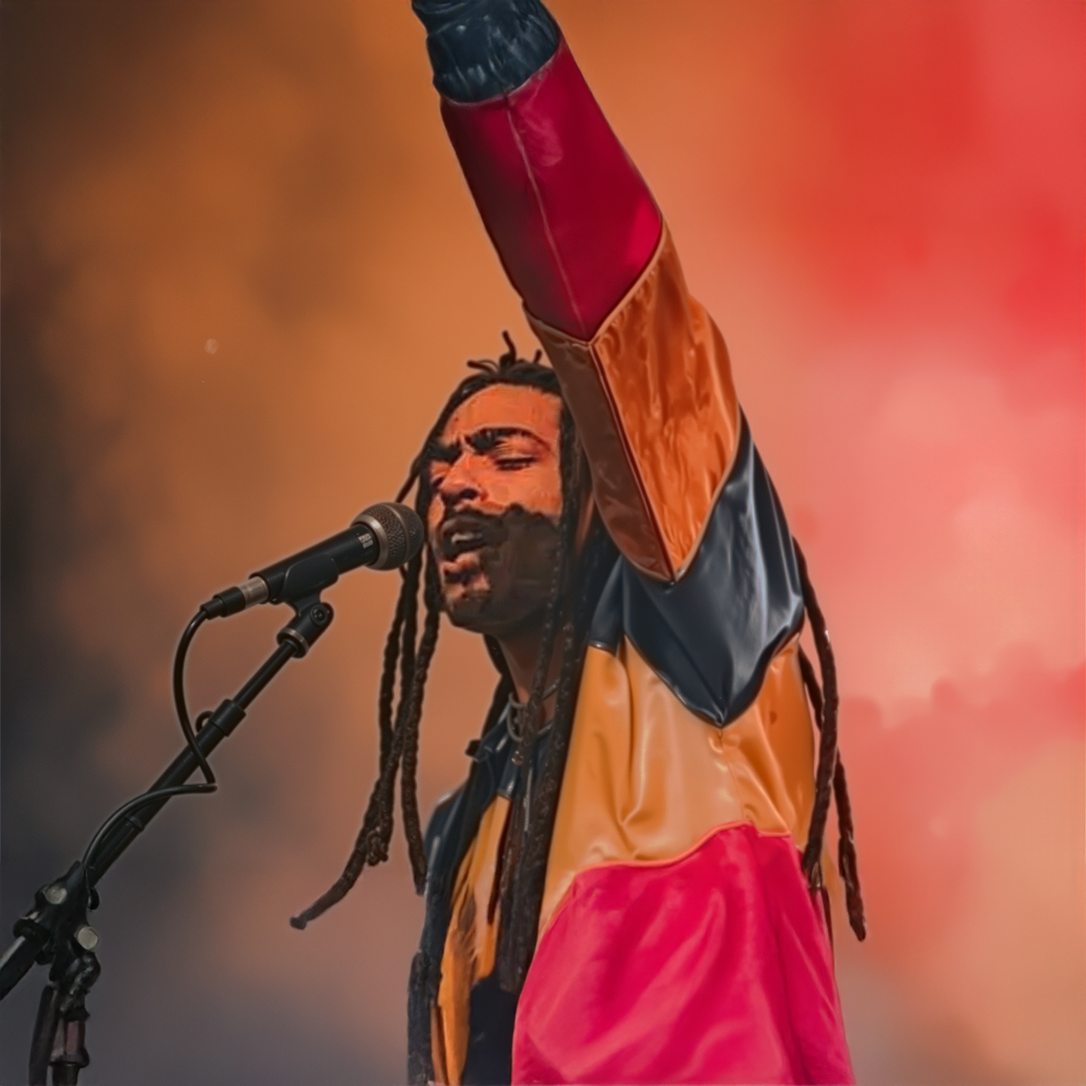 Performer singing into a microphone on stage with an orange and pink background, wearing a colorful jacket with dreadlocks.