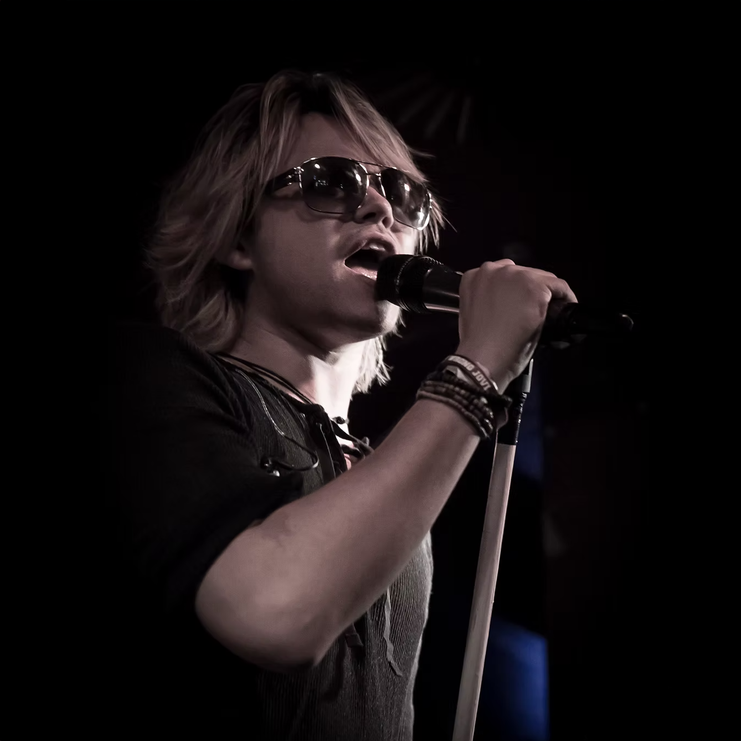 A young man with long blonde hair singing into a microphone on stage, wearing sunglasses and bracelets.