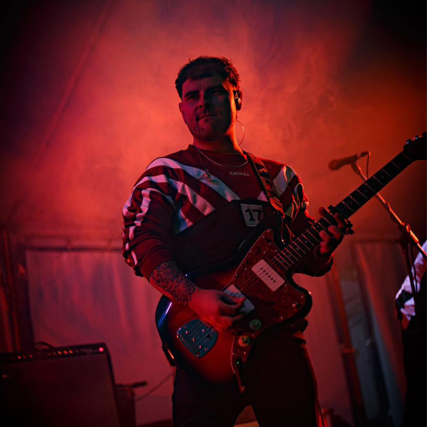 A young man playing electric guitar on stage, surrounded by red and purple lighting, with a microphone and amplifier visible in the background.