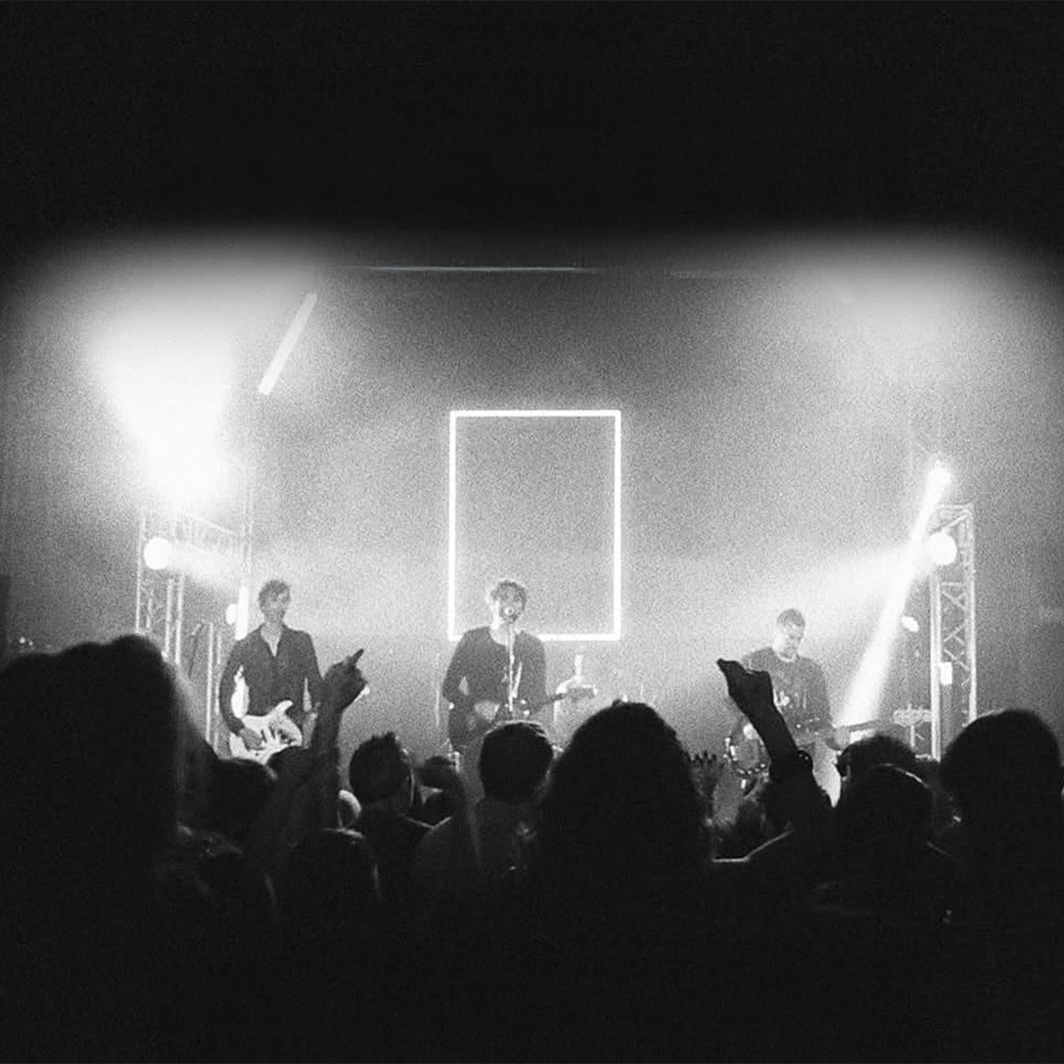 Black and white photo of a live concert with three band members on stage playing guitars, surrounded by bright lights and a crowd in front.