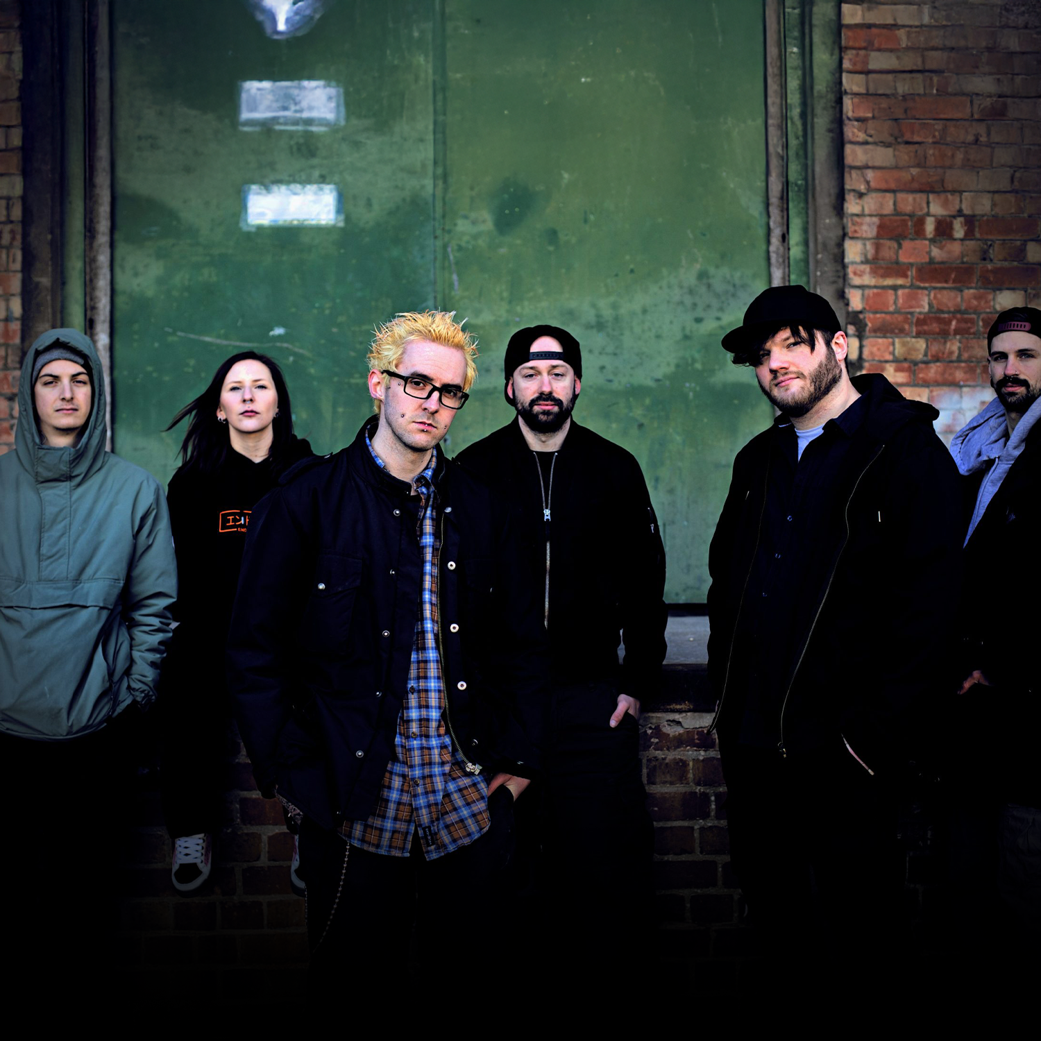 Group of six young adults posing outdoors in front of a worn green wall and brick wall, dressed in casual streetwear.