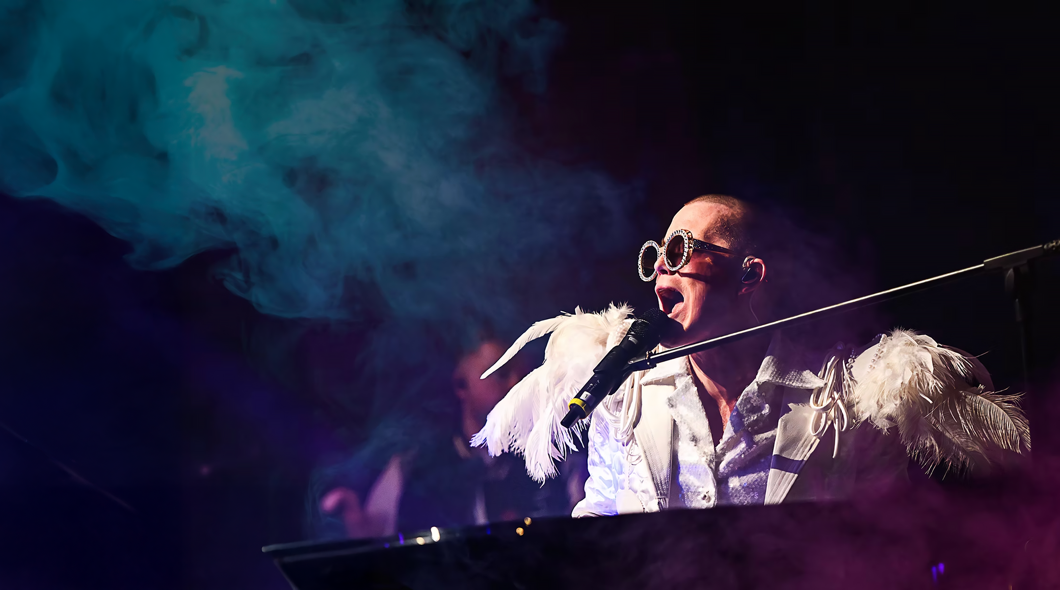 Performer singing into a microphone on stage with colorful haze effects, wearing large novelty sunglasses and a costume with feathers.