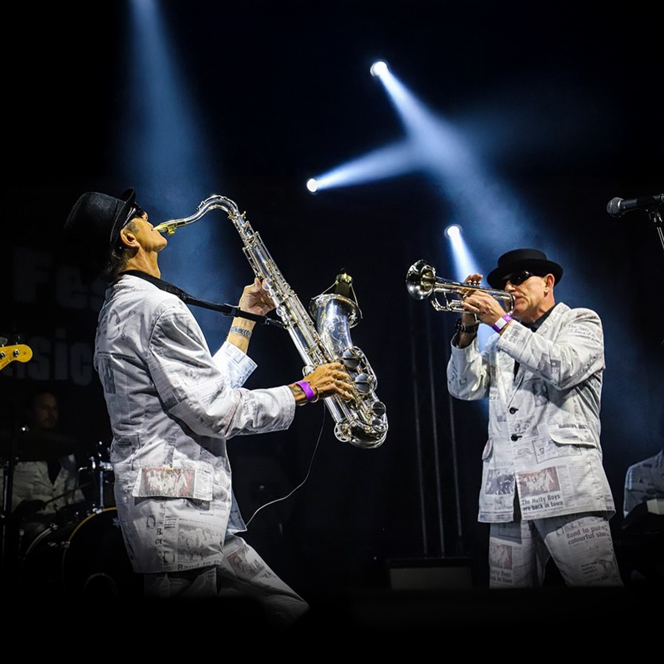Two musicians in black hats and newspaper-print suits playing saxophone and trumpet on stage under blue spotlights.