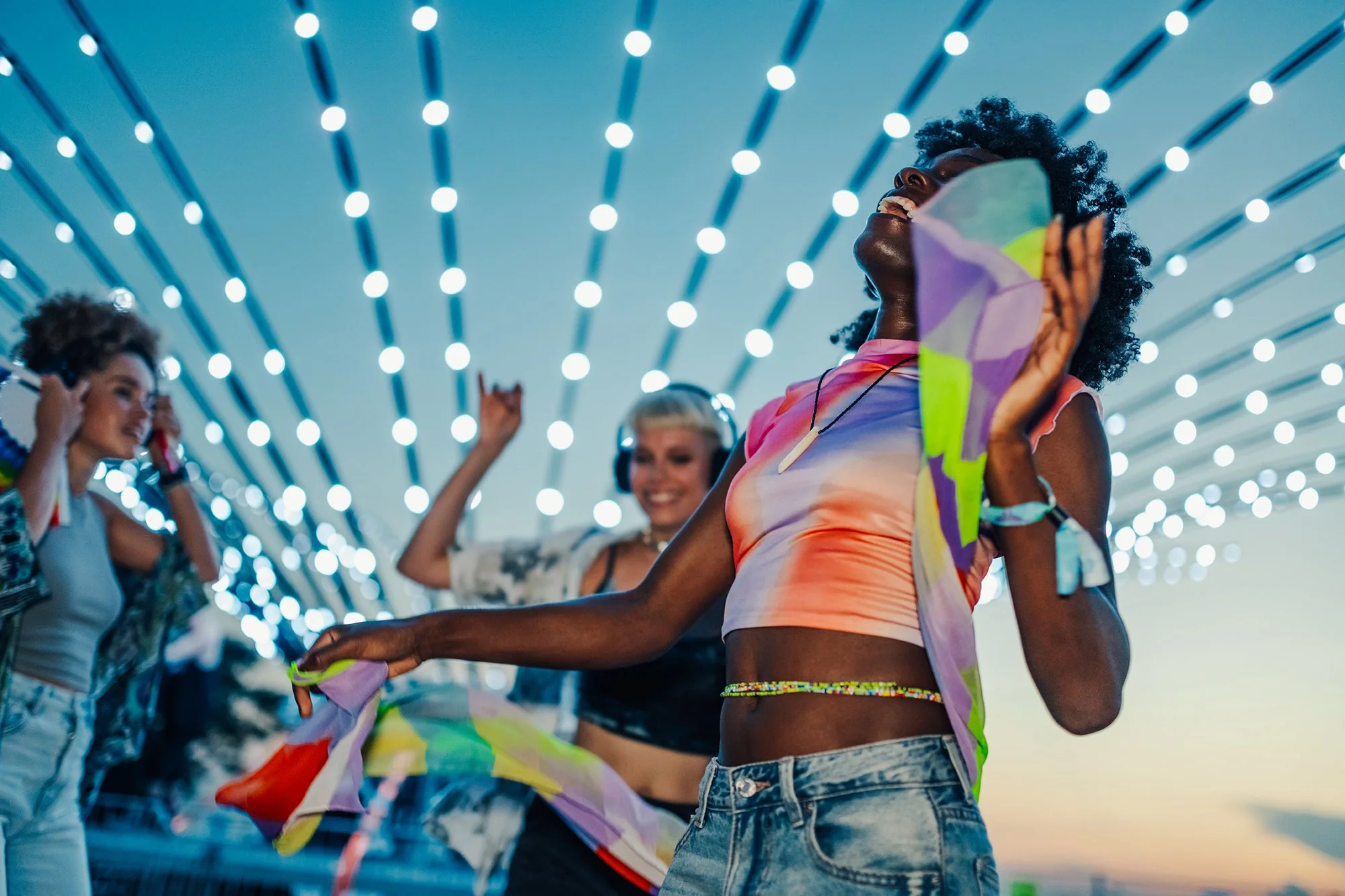 Group of young women dancing and enjoying music at an outdoor event with string lights overhead during sunset