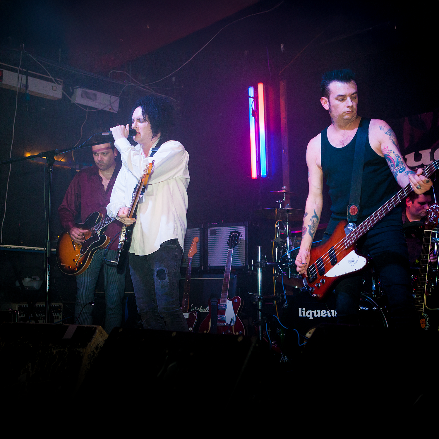 Band performing live on stage with neon lights, featuring a singer with black hair and a guitar, and a bassist with tattoos in a black tank top.