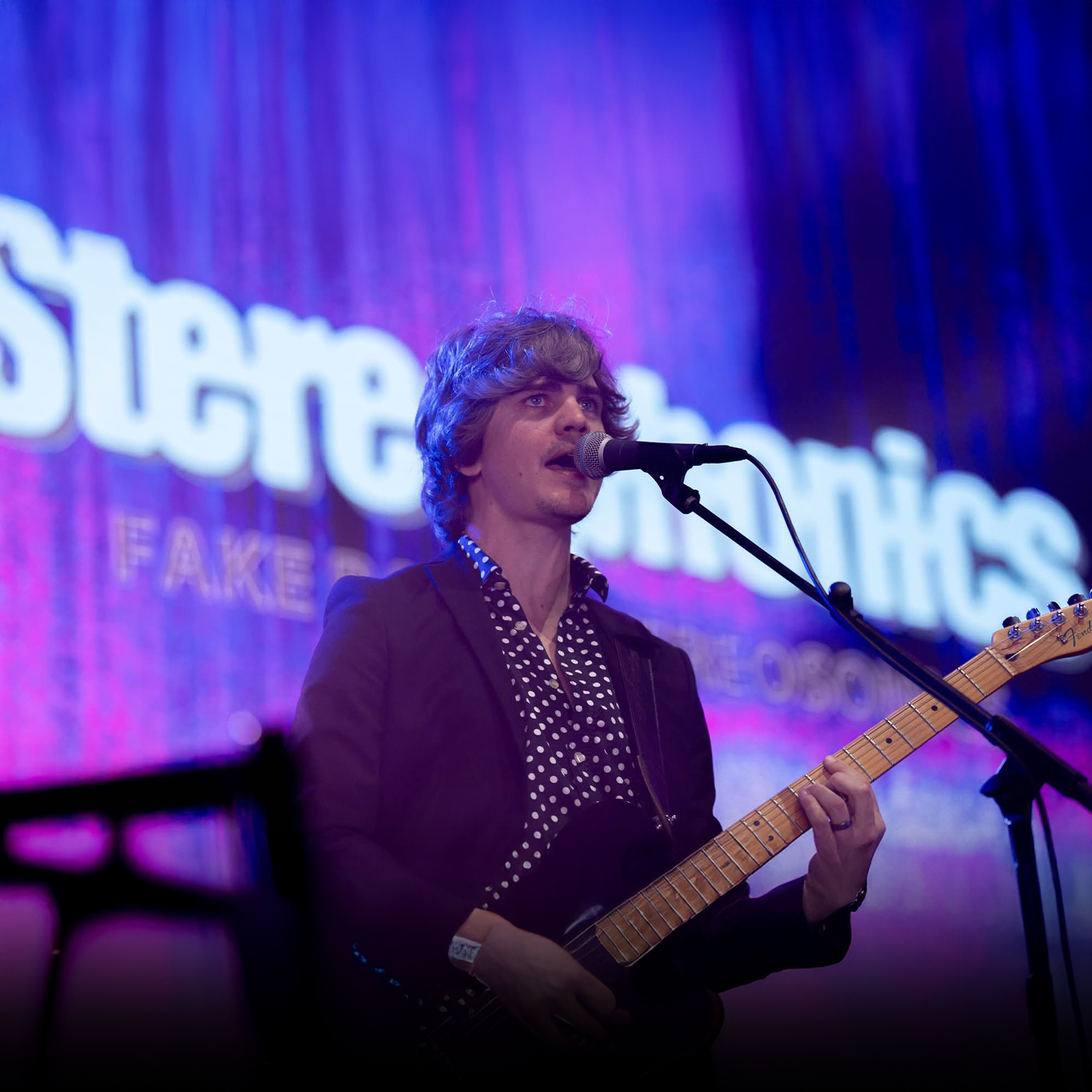 A man with curly hair wearing a black blazer and polka dot shirt, singing into a microphone, playing an electric guitar on stage with a purple and blue background that says 'SUGARY'.