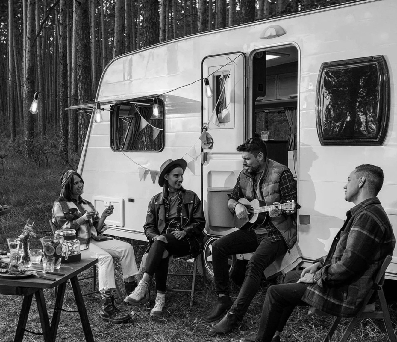 A group of five friends gathered outside near a camper trailer, sitting and standing around a small table and a man playing guitar, with string lights hanging above, surrounded by a wooded forest.