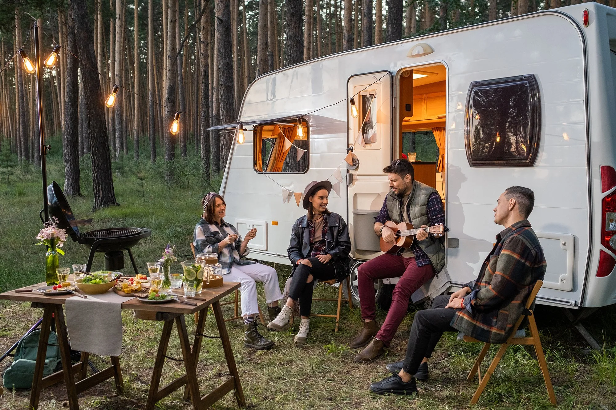 Group of friends enjoying a gathering outside a camper truck in the woods, with a table of food and a person playing guitar.