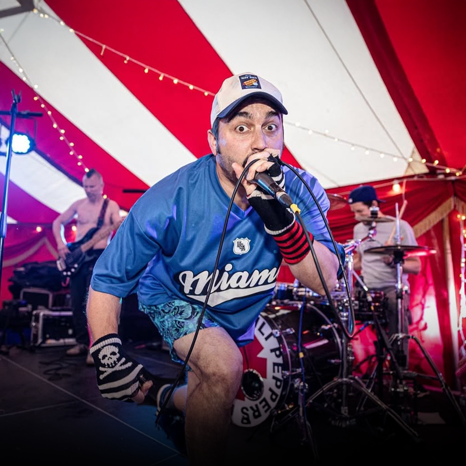 A man in a blue Miami sports jersey and a baseball cap passionately singing into a microphone during a live band performance inside a red and white striped tent. Two other band members are seen in the background playing instruments.