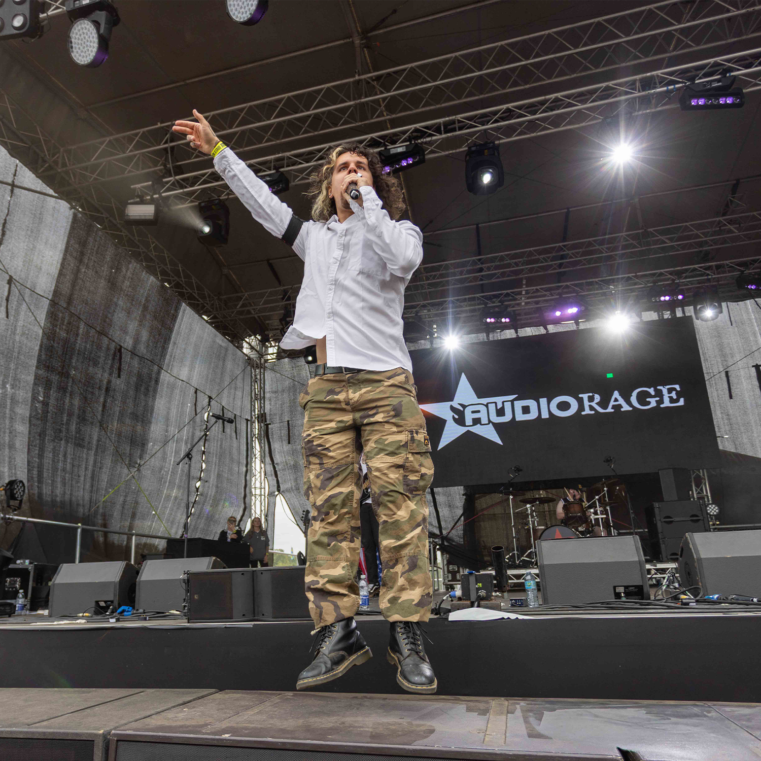 A male performer with long curly hair, wearing a white button-up shirt, camouflage pants, and black boots, singing into a microphone on a stage at a music event. The stage features a large screen displaying the "Audiorage" logo, and stage lights are shining brightly.