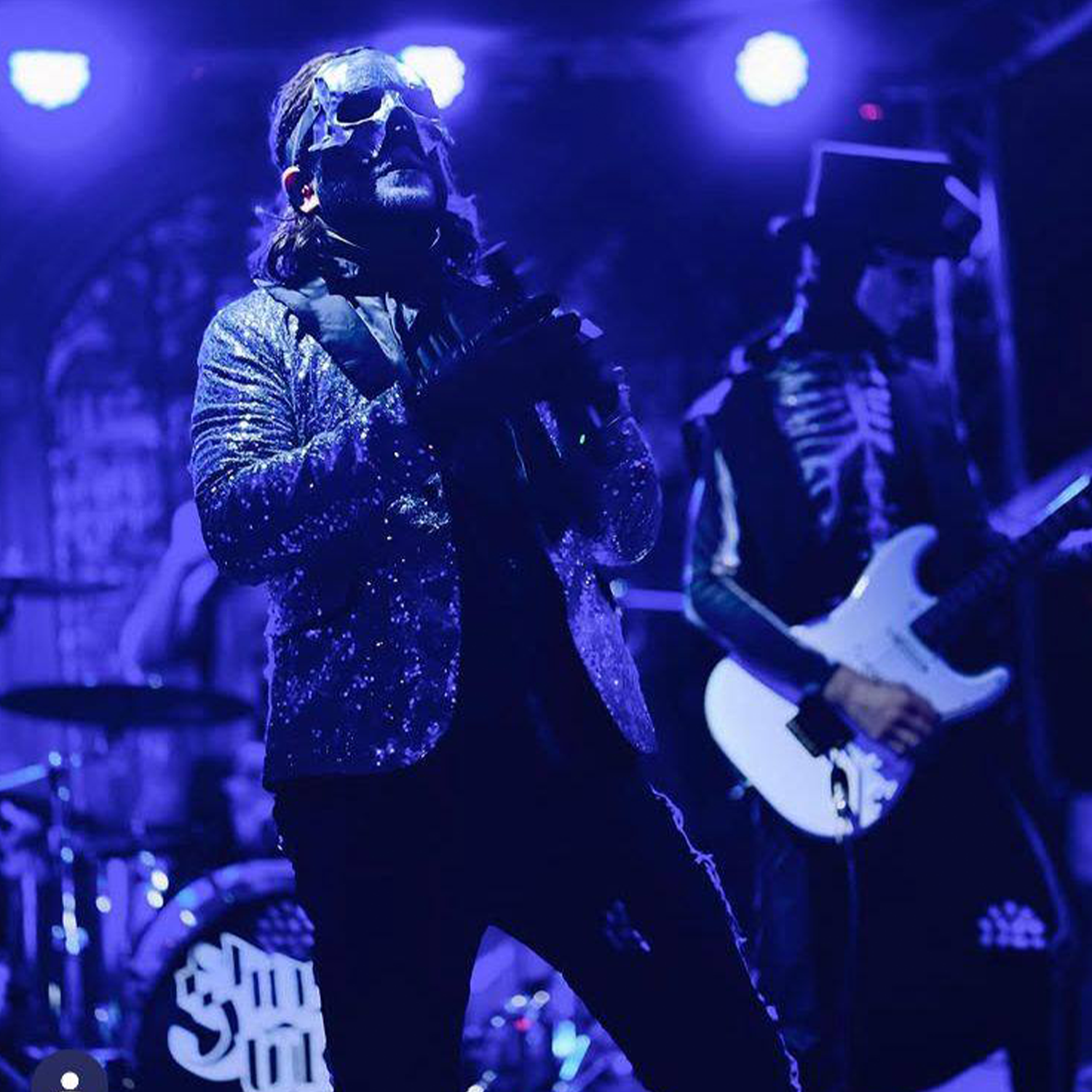 Musician wearing a shiny, starry blazer and sunglasses, singing into a microphone on stage under purple lighting, with a guitarist in skeleton costume and a drum set in the background.