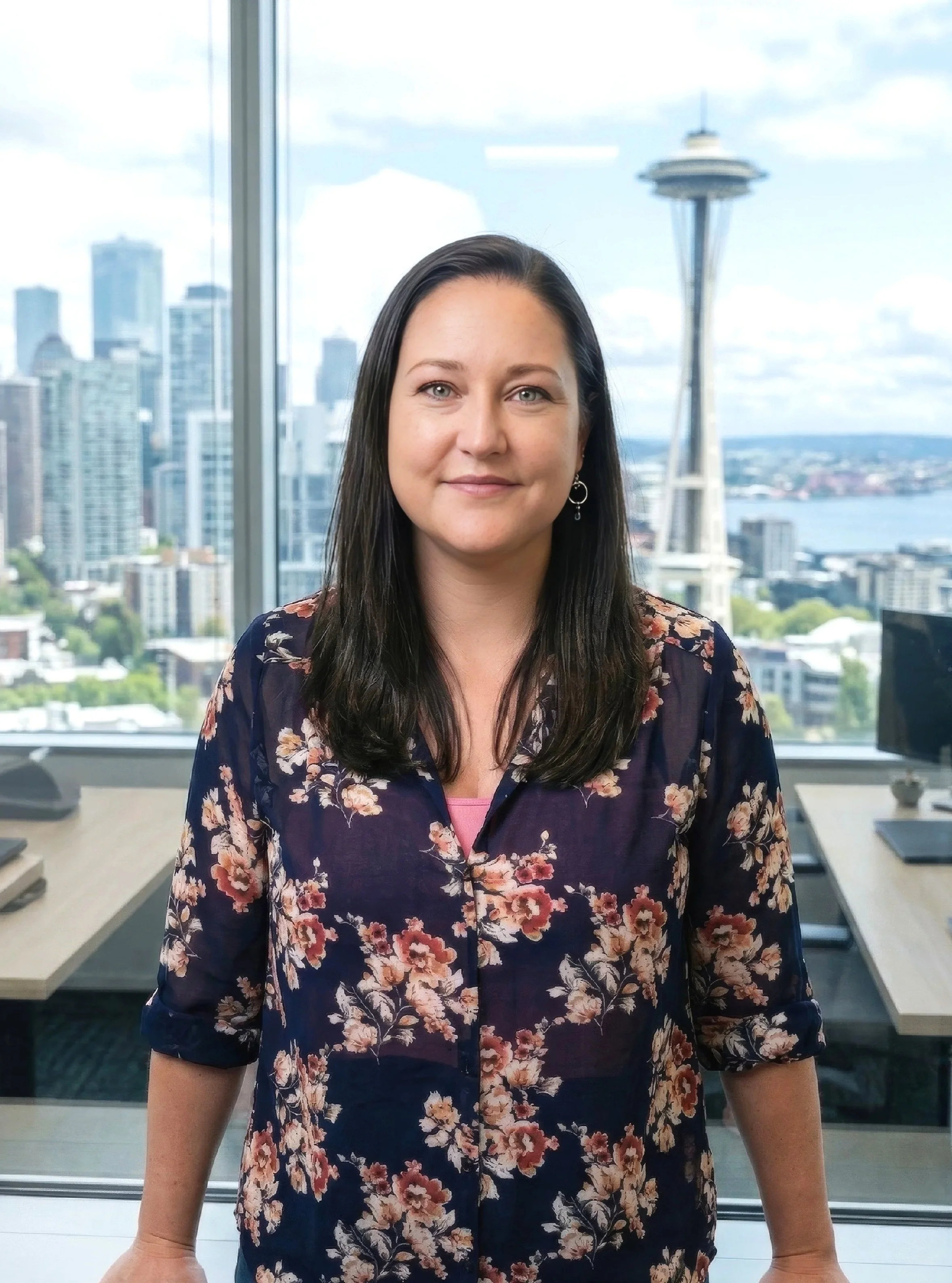 Dawn Moser wearing a floral blouse standing in an office with large windows overlooking Seattle skyline.