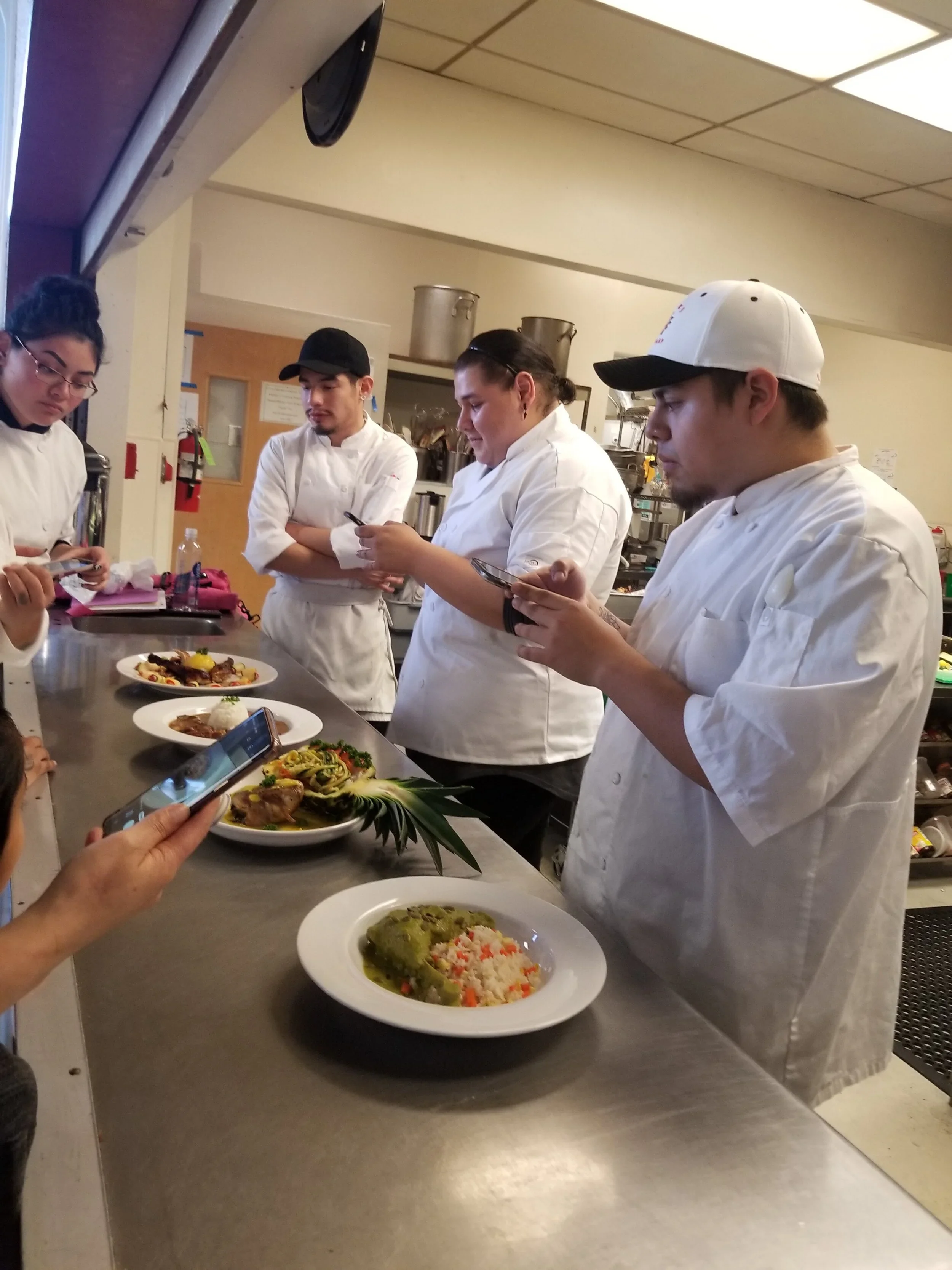 MLVS Culinary Program. Group of four chefs in white uniforms at a kitchen counter taking pictures of plated dishes with phones, with a woman in the foreground also taking a photo.
