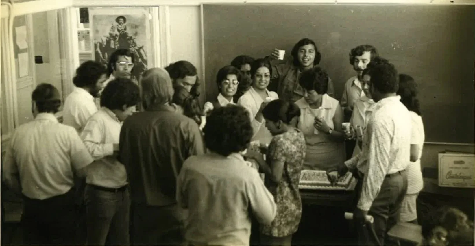 MLVS History: A group of people gathered around a table near a blackboard in a classroom, celebrating a birthday with a cake.