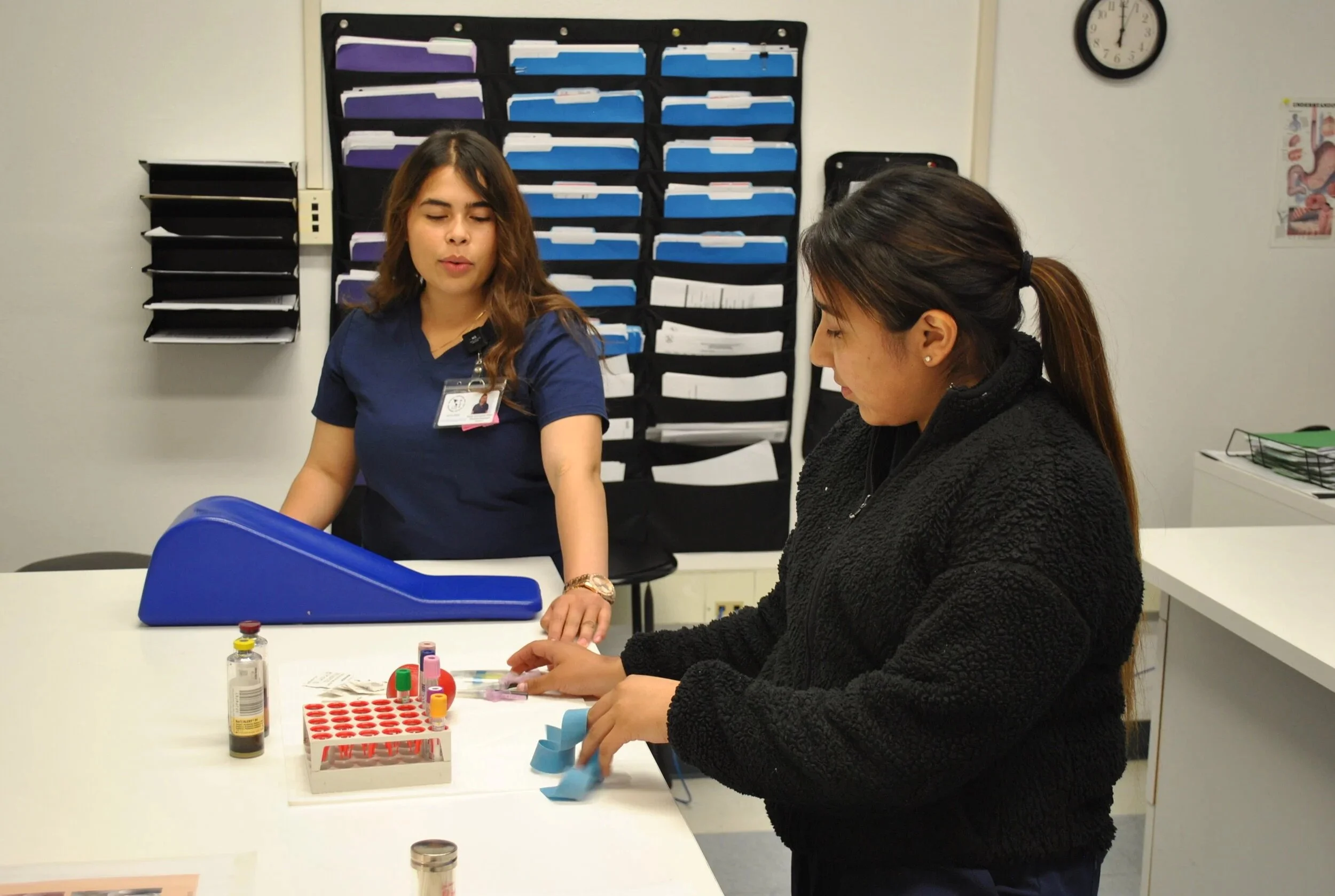 MLVS Medical Assistant Program: Two women in a medical setting, with one woman in a navy scrub top and the other in a black fleece jacket, are handling medical supplies, including vials and test tubes, on a white table.