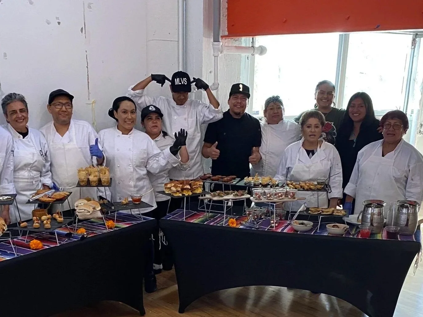 MLVS Culinary Program: Catering. Group of chefs and staff standing behind a table filled with assorted foods and appetizers, inside a bright room with large windows.