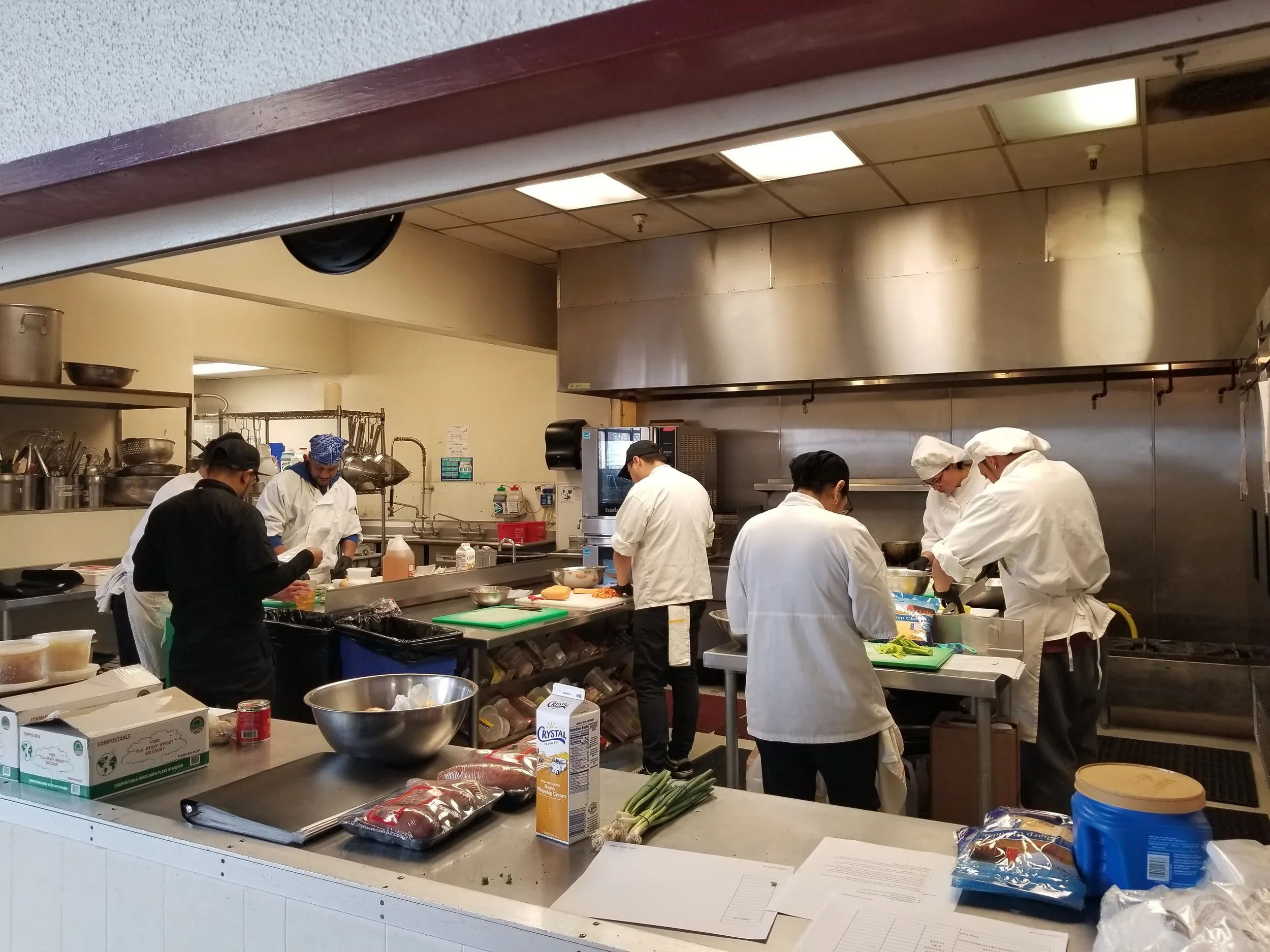 MLVS Culinary Program: Kitchen with six chefs preparing food at a counter, some chopping ingredients, and various kitchen supplies and utensils nearby.