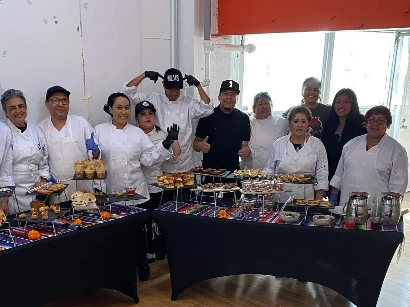 MLVS Culinary Arts Program. Group of chefs and staff standing behind a table with an array of food dishes, some chefs wearing aprons and gloves, one man flexing muscles, another giving a thumbs up, interior with large windows in the background.