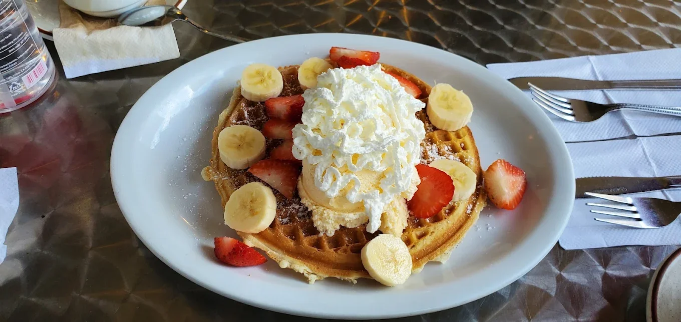 Jims By MLVS: Waffles. A plate with a waffle topped with banana slices, strawberry slices, whipped cream, and powdered sugar.