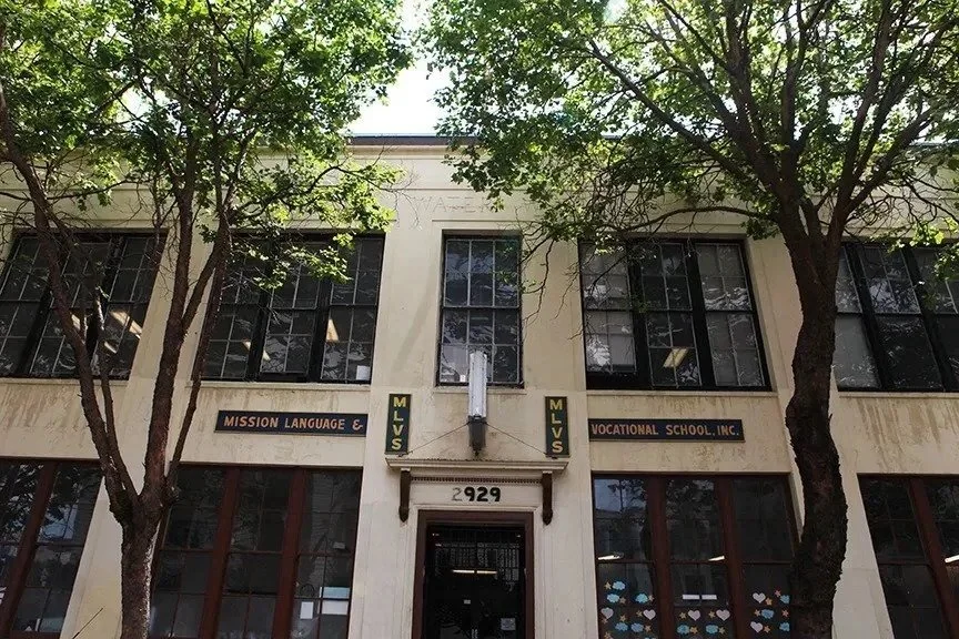 Front view of a two-story building with signs for Mission Language & Vocational School Inc., surrounded by trees, with the address 2929 above the entrance.