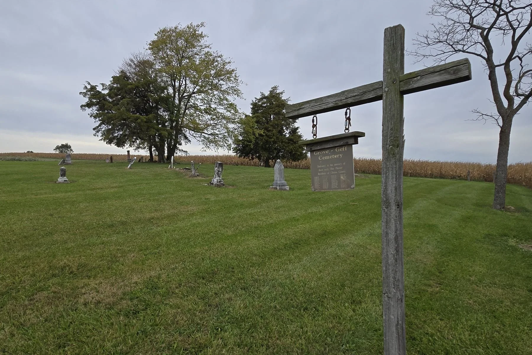 A weathered wooden post holds a hanging sign lettered Grove - Geff Cemetery above columns of names and years, with headstones, trees and a cornfield behind.