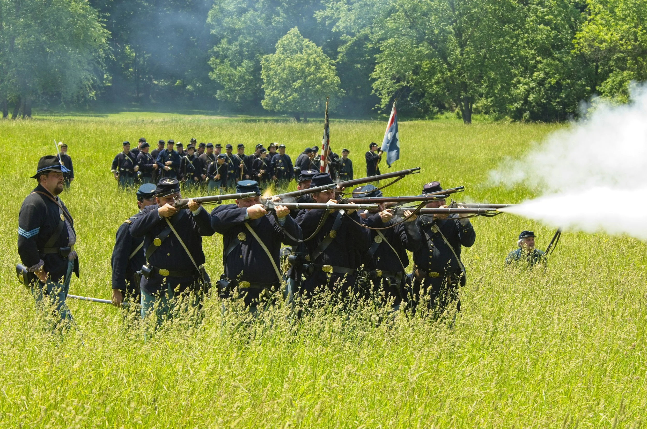 Civil war reenactors dressed in blue uniforms firing muskets in a grassy field with trees in the background.
