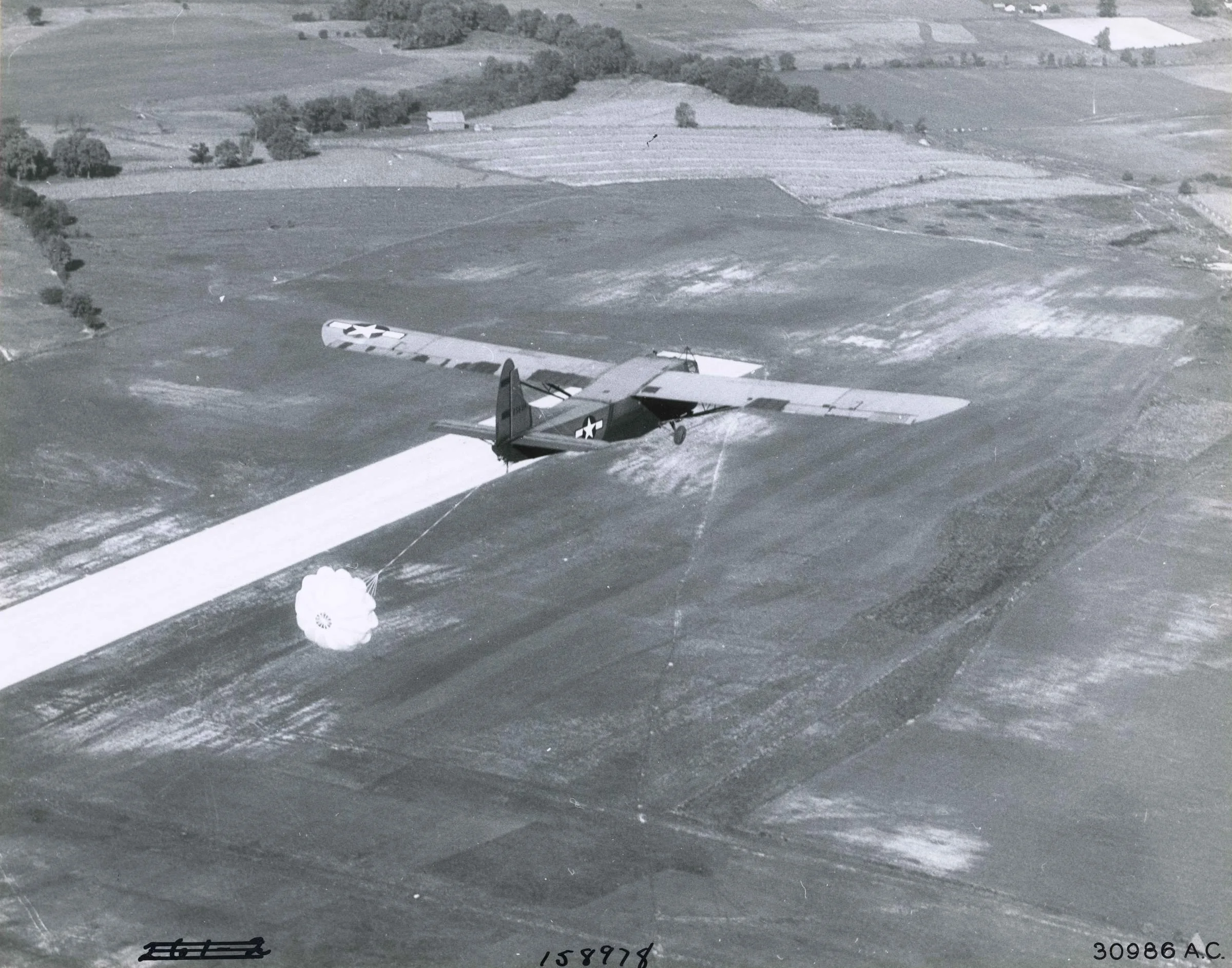1944 aerial photograph of a Waco CG-4A glider descending over the airfield runway, with the cleared orchard tract at upper right.