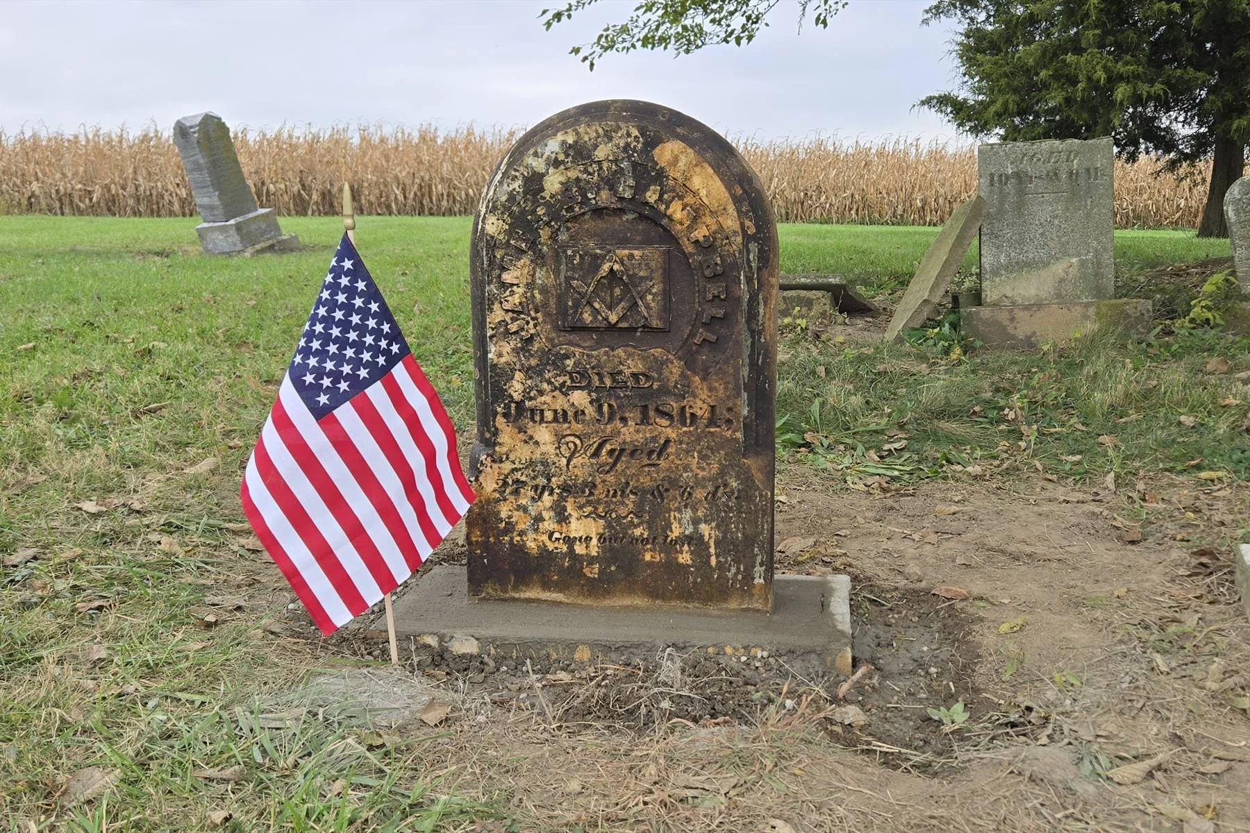 An arched marble headstone, darkened with age, stands upright on a fresh concrete base beside a small American flag, with older stones and a cornfield behind it.