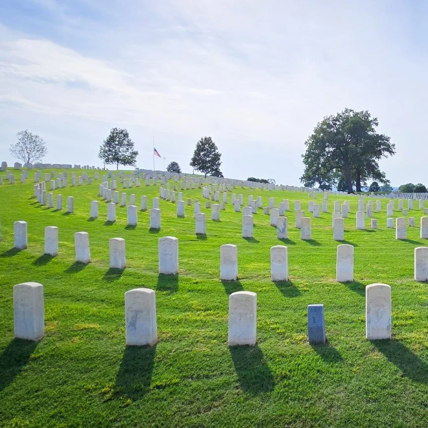 A military cemetery with white headstones on green grass, an American flag, and trees in the background under a partly cloudy sky.